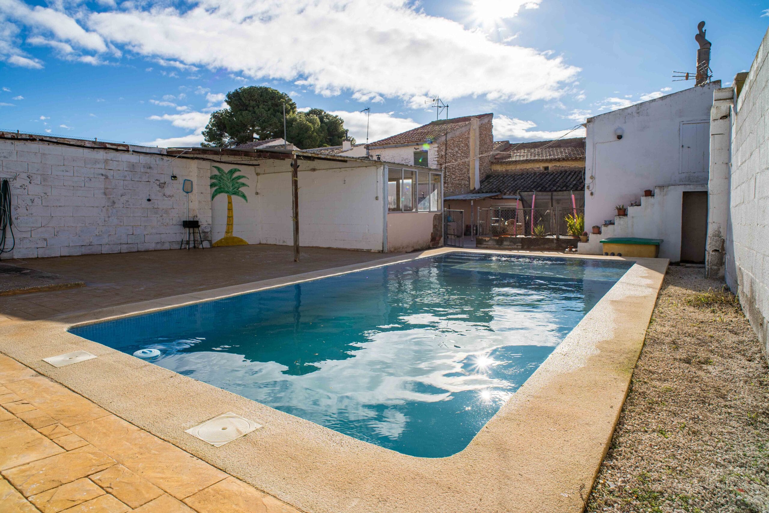 A private outdoor swimming pool with clear water, surrounded by paved and gravel surfaces, enclosed by white walls and nearby residential structures under a partly cloudy sky.