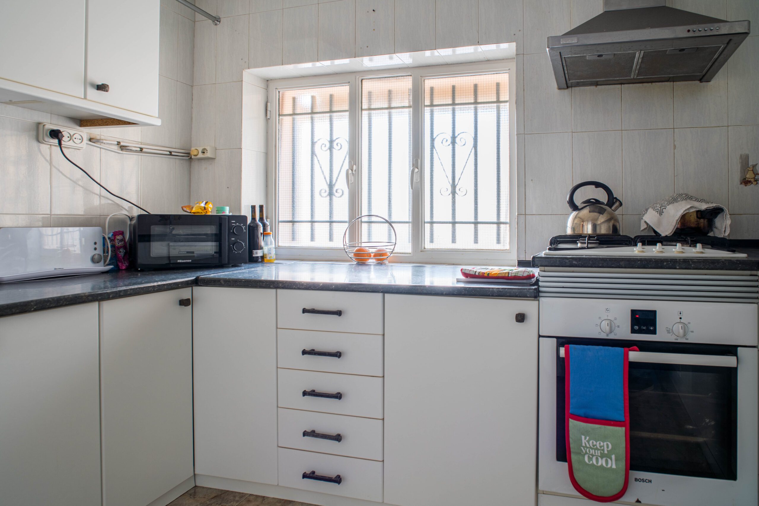 Compact kitchen featuring white cabinets, a black countertop, a window with security bars, a microwave, stove with kettle, and an oven with colorful towels hanging.