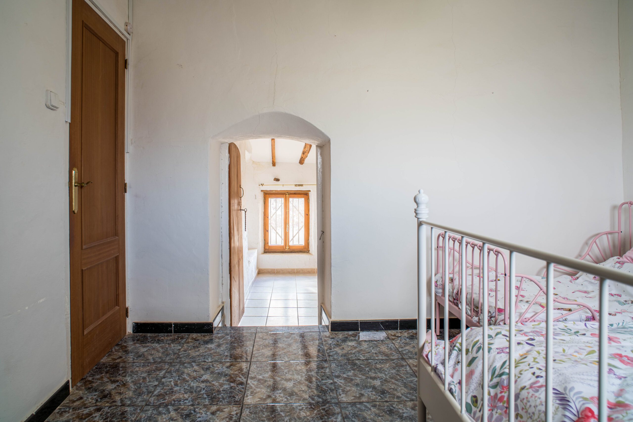 A child's bedroom with a pink metal bed frame and floral bedding, tiled floor, white walls, and a wooden door leading to another room with a window.
