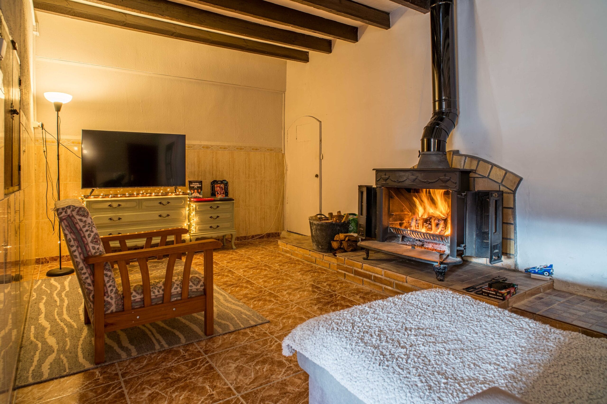 A cozy living room with a burning wood stove fireplace, wooden armchair with patterned cushions, a TV on a light-colored dresser decorated with string lights, and exposed wooden ceiling beams.