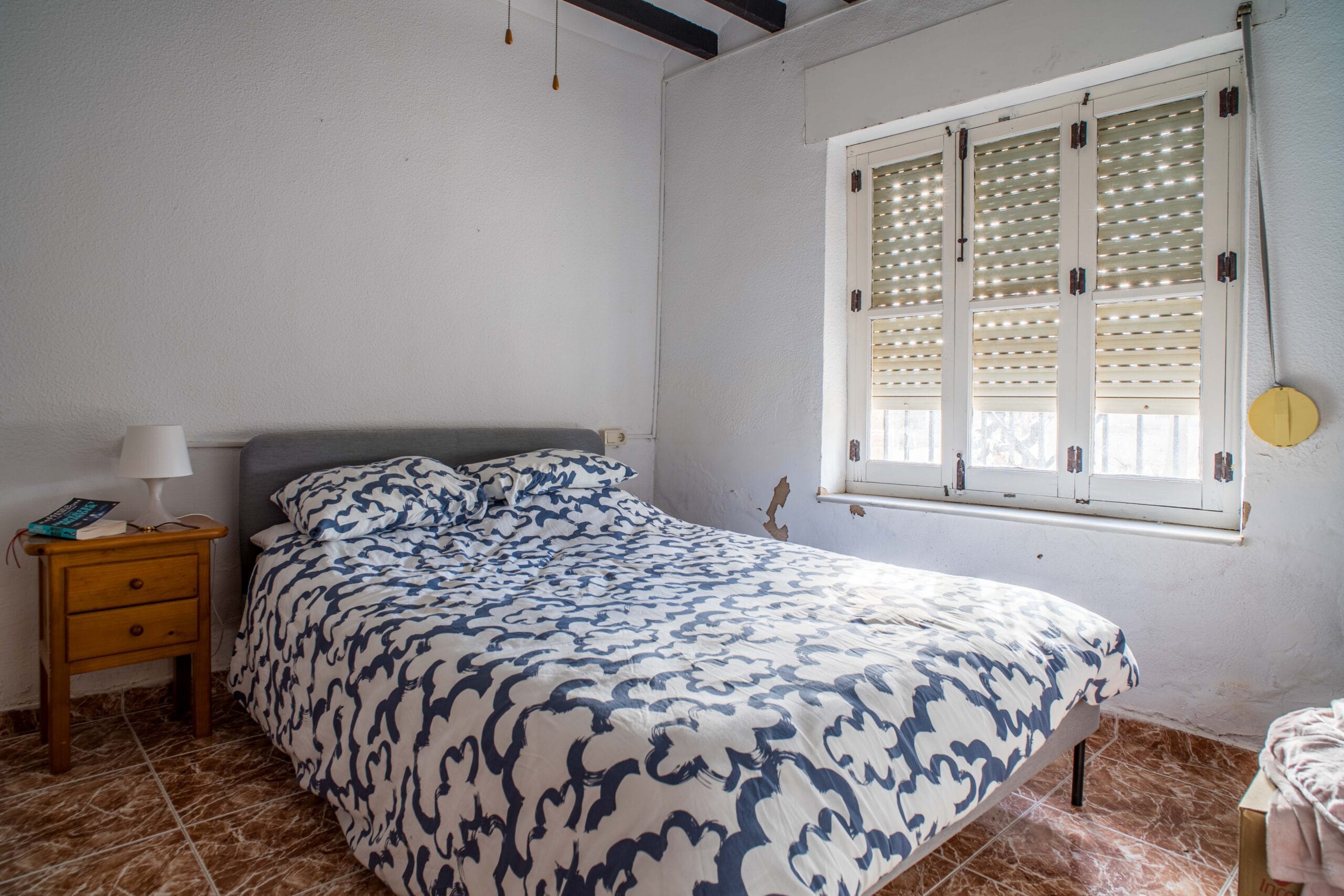 A bedroom featuring a double bed with patterned bedding, a wooden bedside table with a lamp and books, and a window with partially closed shutters letting in light.