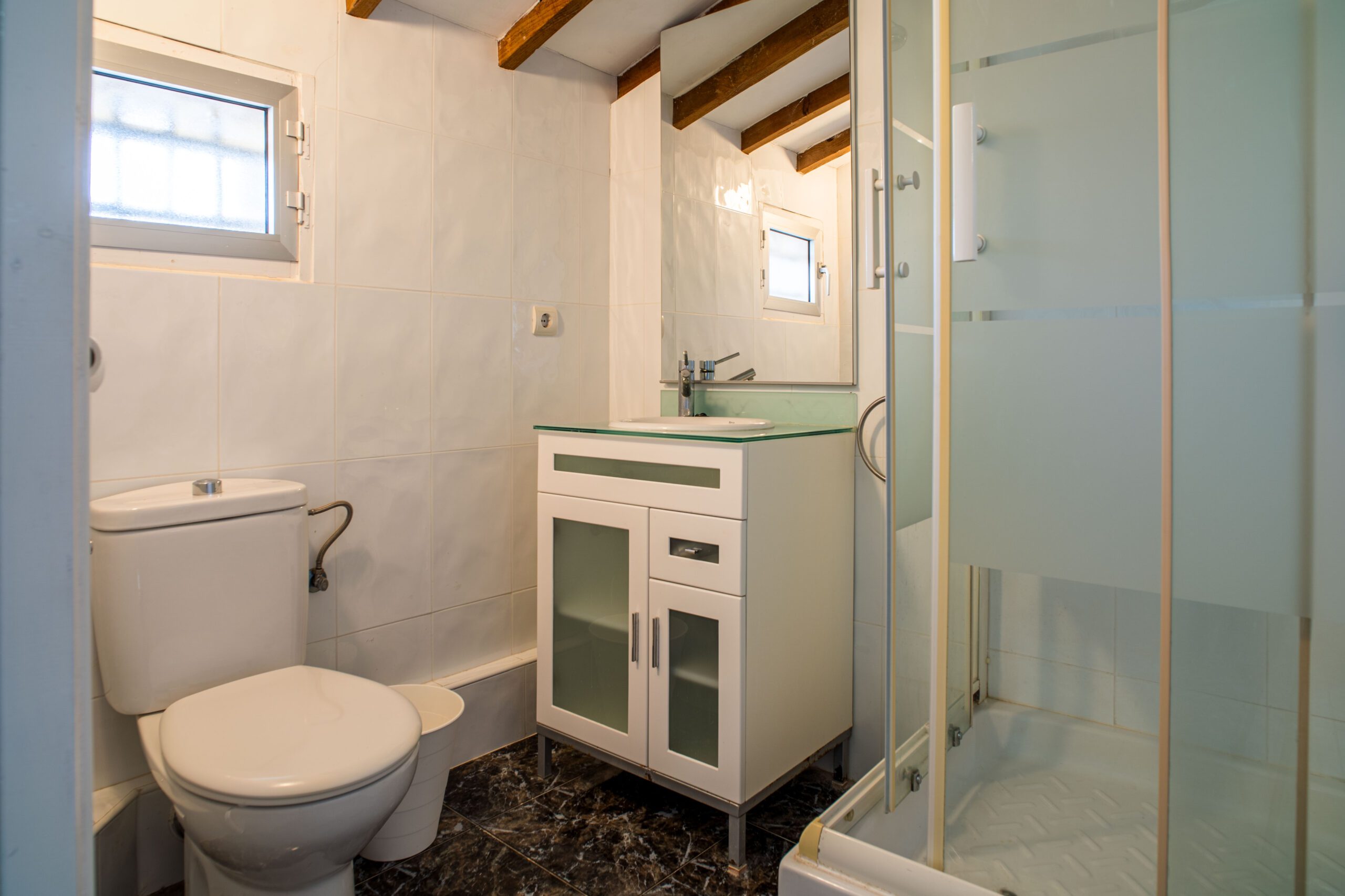 Small bathroom with white tiled walls, a toilet, a white vanity with a glass countertop and sink, and a shower with frosted glass doors.
