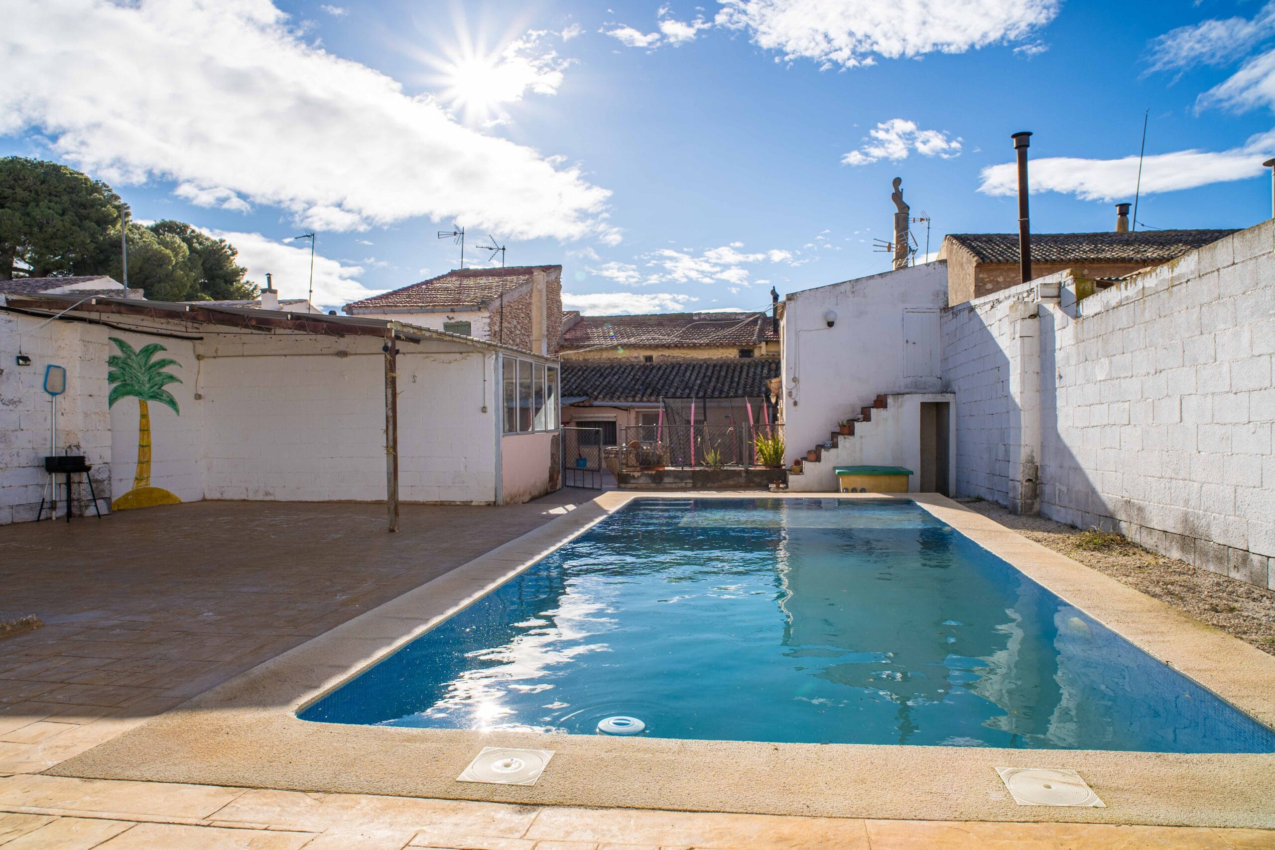 Outdoor swimming pool with clear blue water, surrounded by a tiled terrace and white walls, under a bright sunny sky with some clouds.