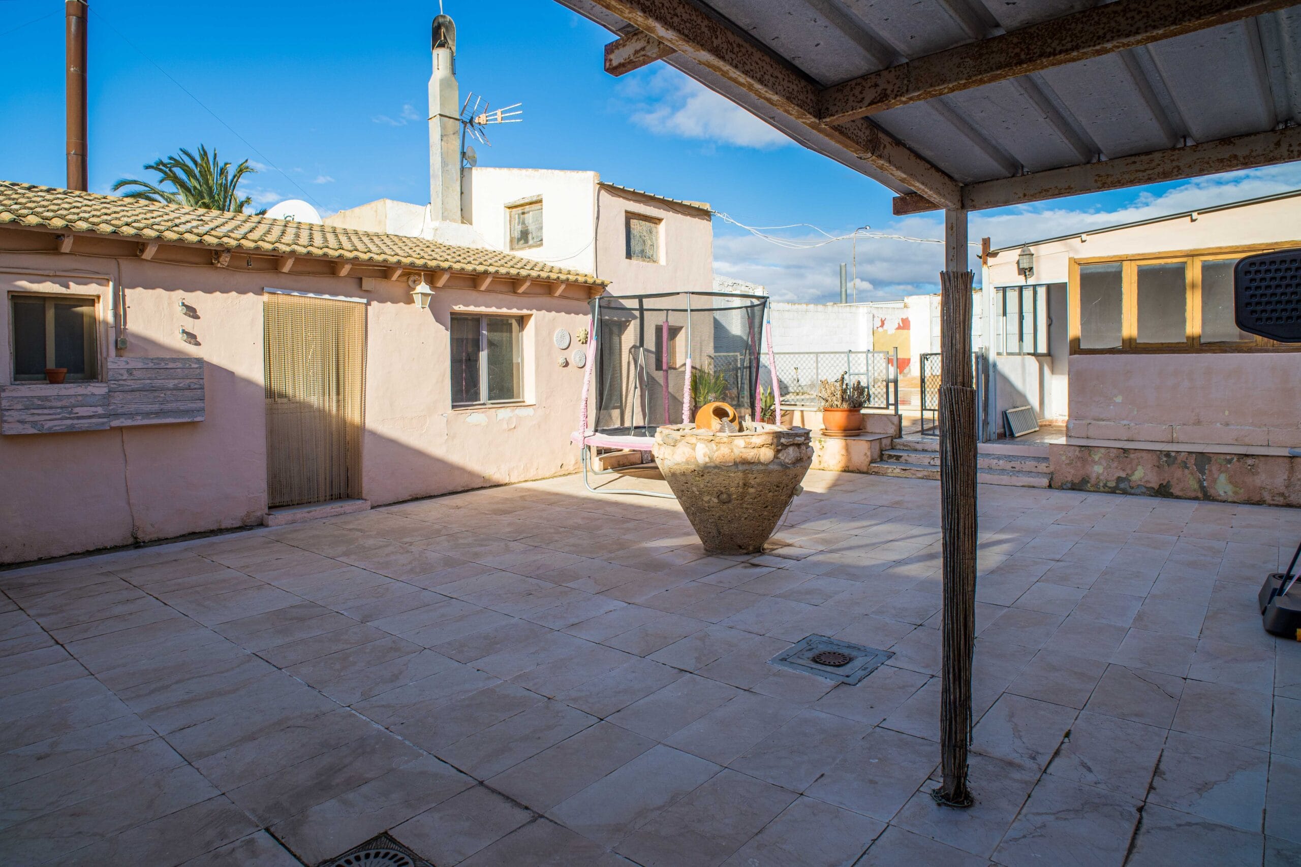 Outdoor courtyard with tiled floor, rustic pink walls, a covered metal roof area, and a clear blue sky.