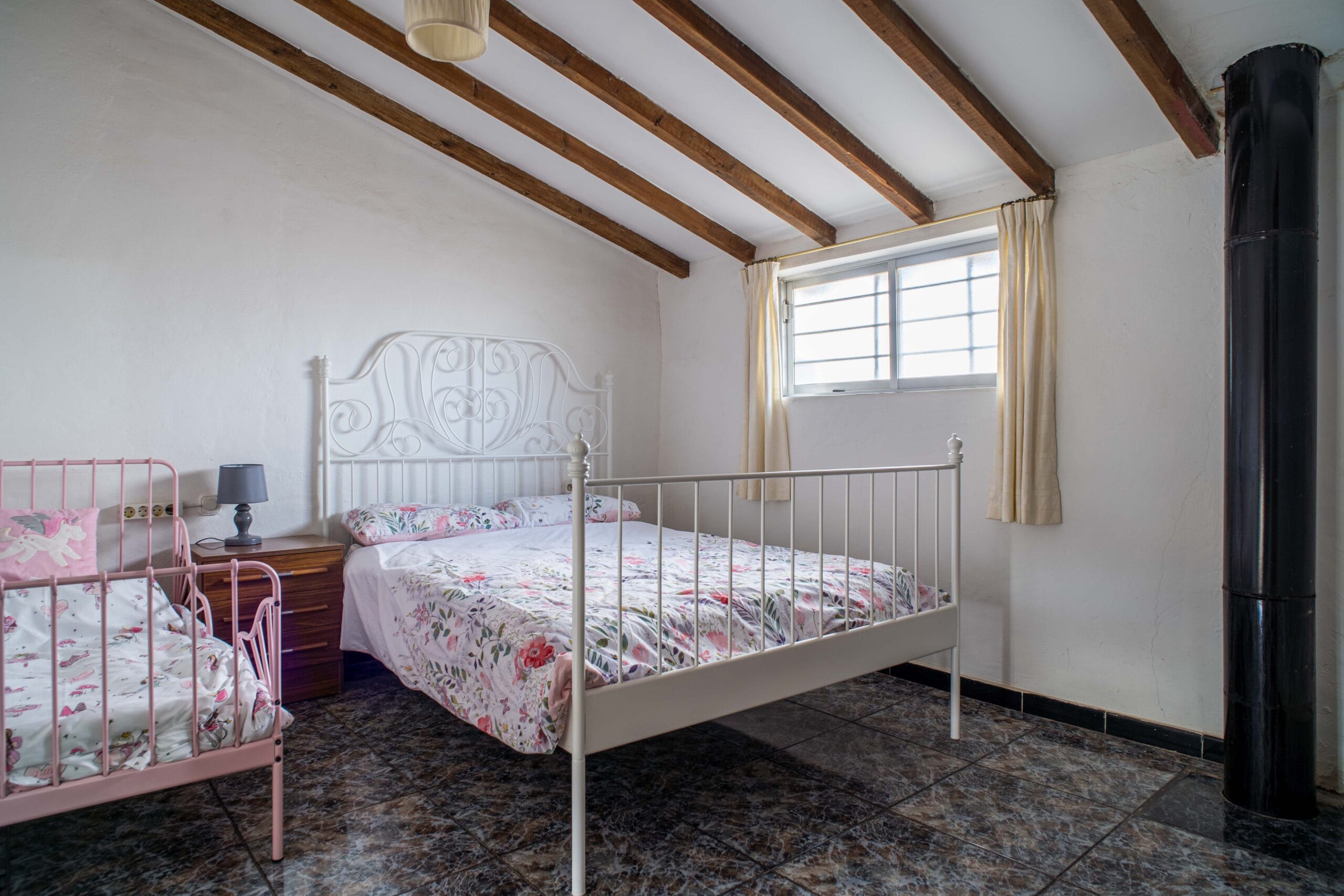 Bedroom with a white metal double bed and a pink metal twin bed, floral bedding, exposed wooden ceiling beams, a window with cream curtains, a bedside table with a lamp, and a black cylindrical stove pipe.