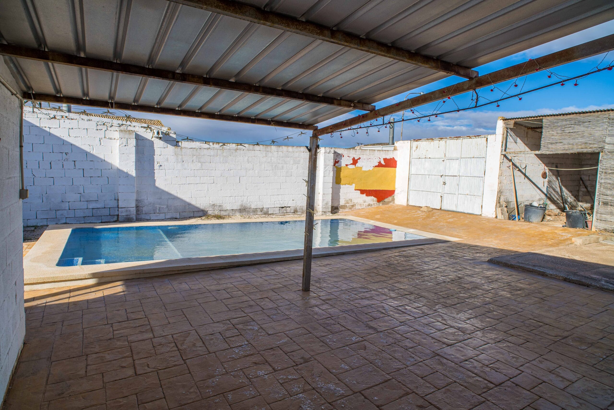 View of a covered patio with a metal roof and a small private swimming pool surrounded by white brick walls under a blue sky.