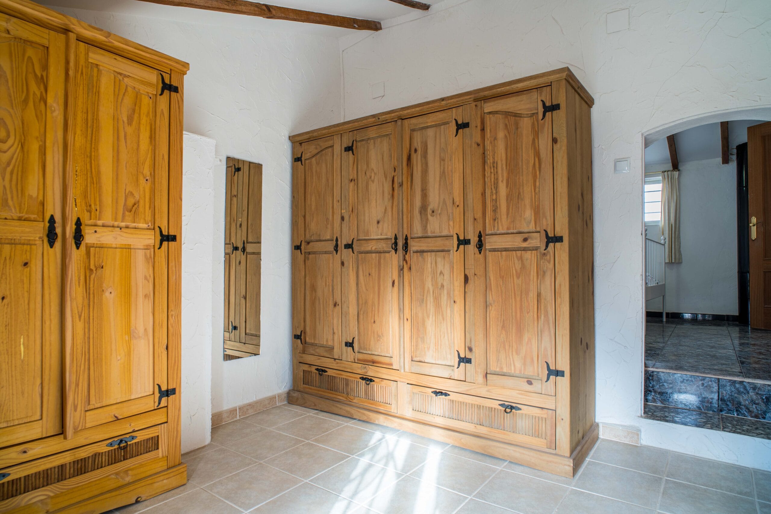 Interior view of a storage or dressing area with multiple large wooden wardrobes, a full-length mirror on a white textured wall, and tiled floor illuminated by natural light.