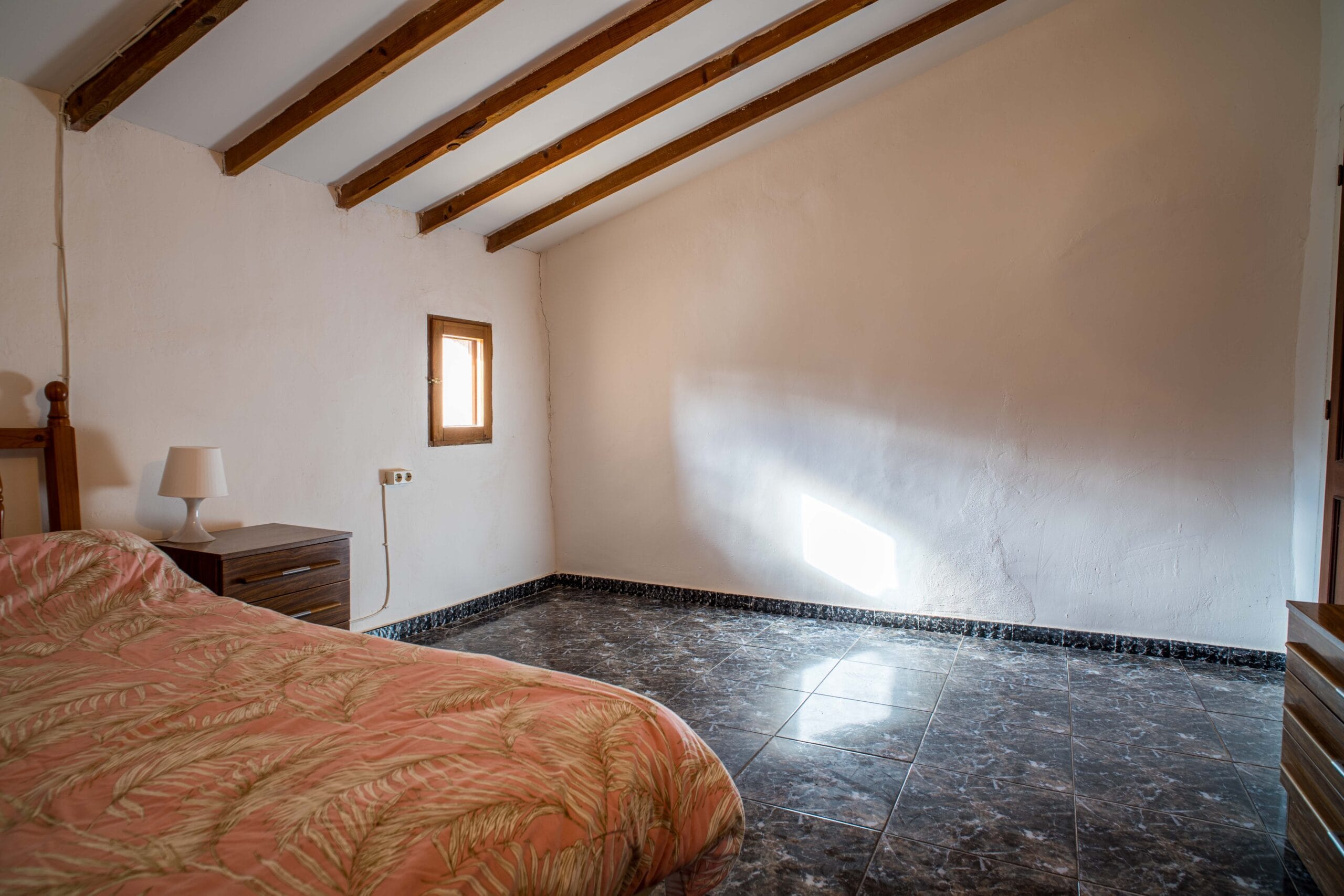 A bedroom featuring a wooden bed with a patterned bedspread, a bedside table with a white lamp, small window, and exposed wooden ceiling beams.