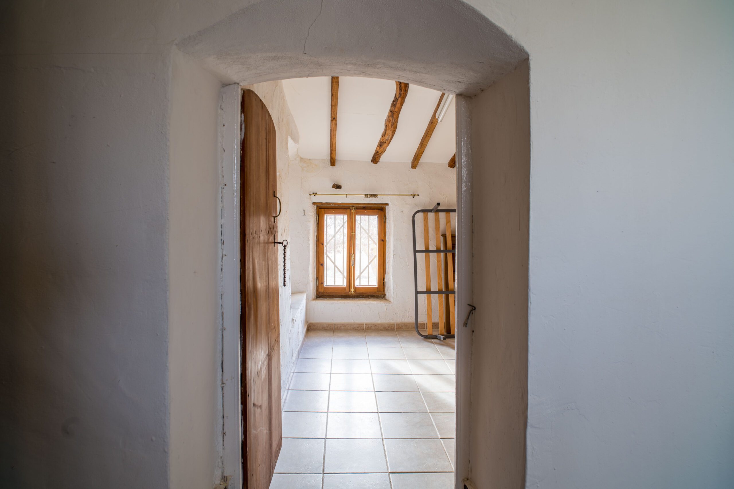 Interior view through an arched doorway into a small room with tiled floor, wooden window with metal bars, and exposed wooden ceiling beams.