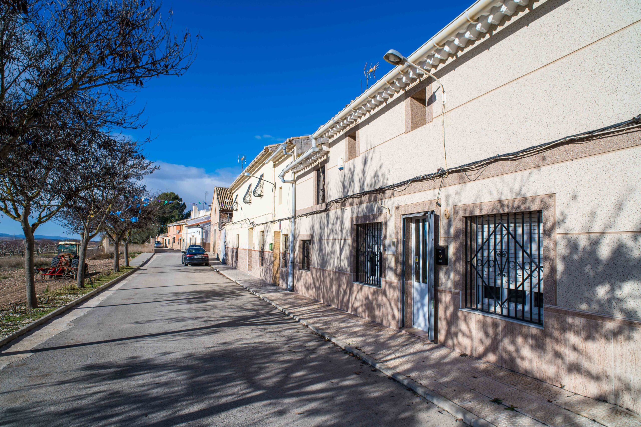 A street lined with residential houses featuring gated windows and doors, parked cars along the road, and leafless trees casting shadows on the pavement under a bright blue sky.
