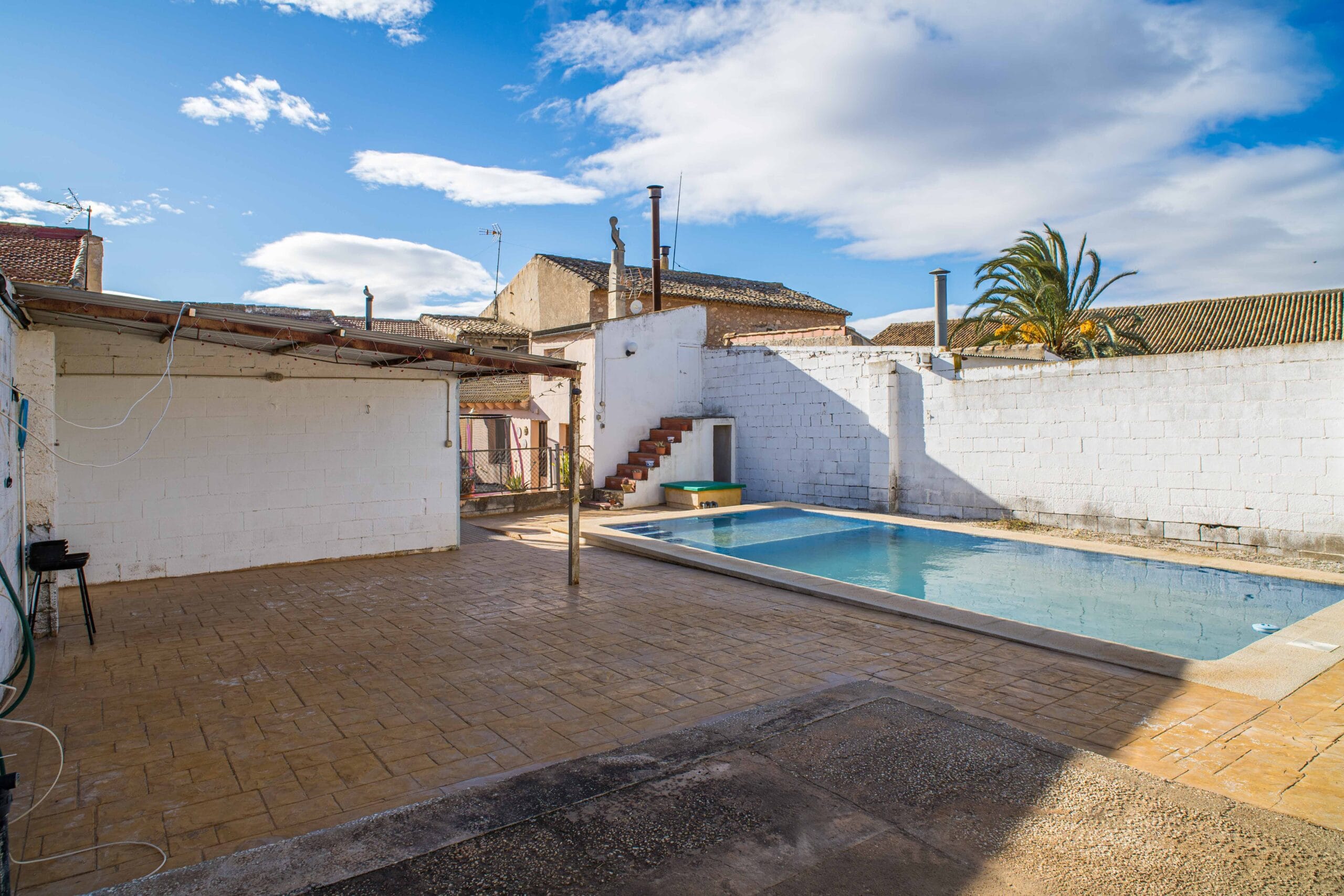 Outdoor private swimming pool with a tiled terrace and white brick walls, under a partly cloudy blue sky.