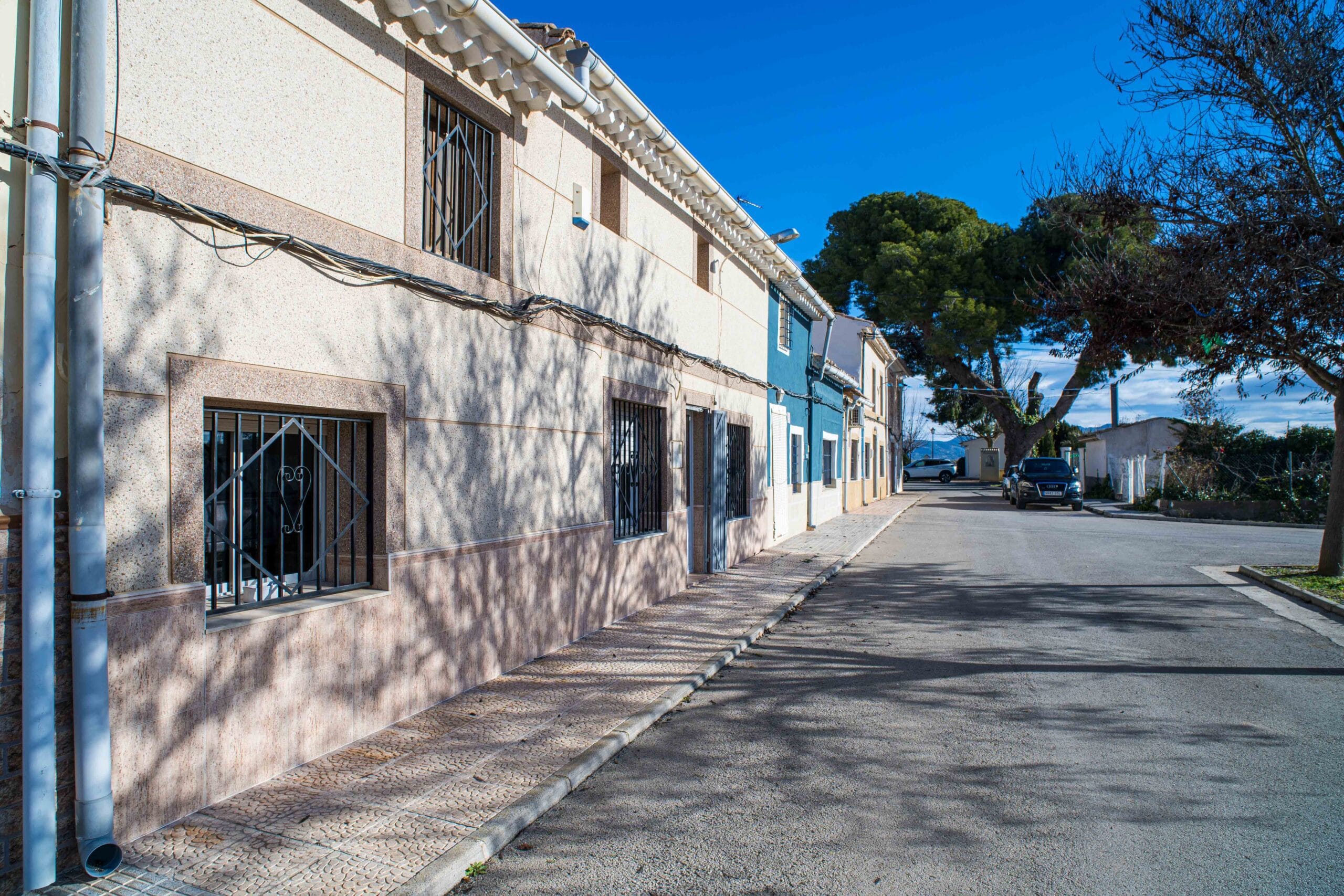 Exterior view of a residential building along a street with barred windows, a sidewalk, parked cars, and trees under a clear blue sky.