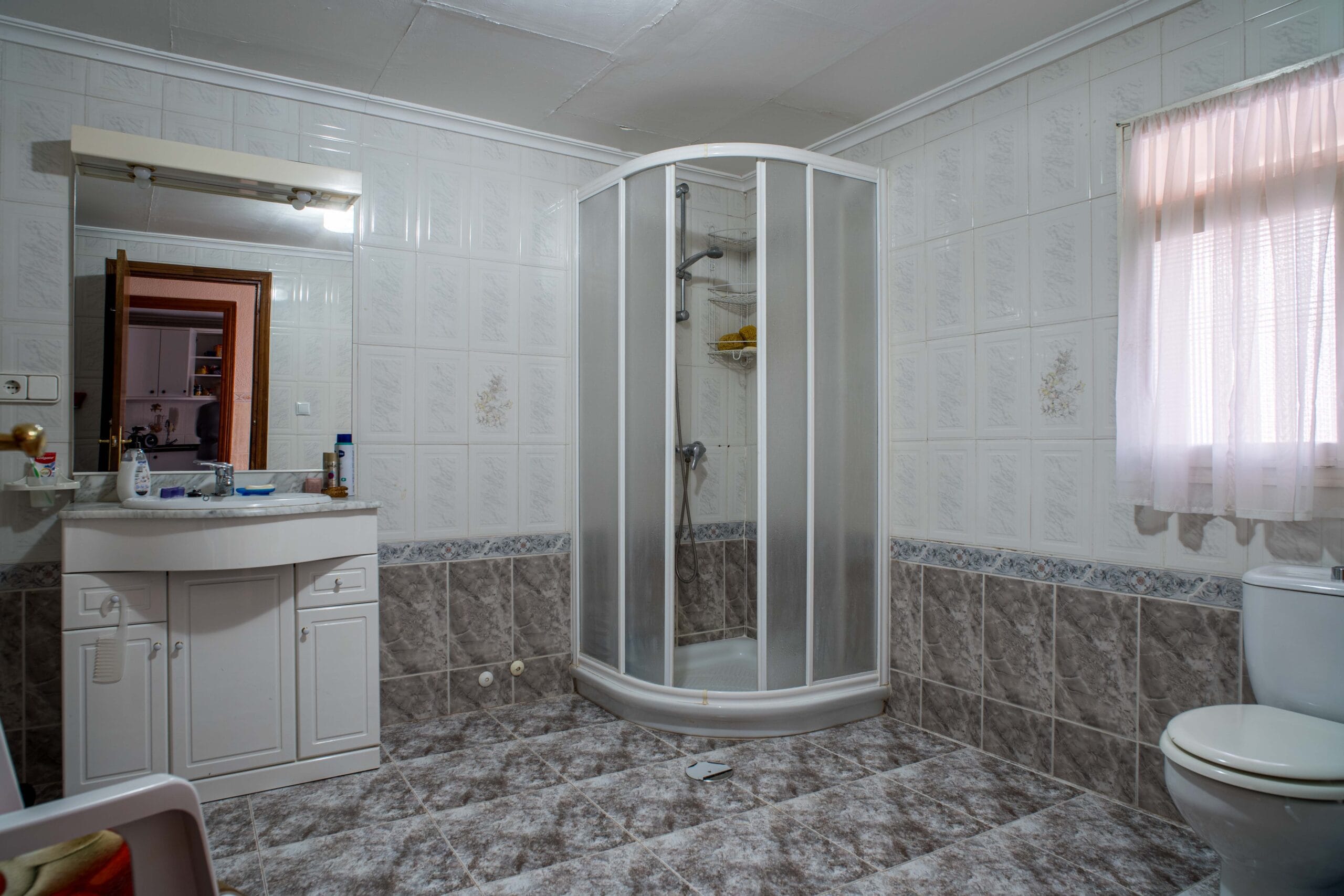Bathroom with a corner shower cabin, white tiled walls with floral accents, grey tiled floor, a vanity with a mirror, and a window with sheer curtains.
