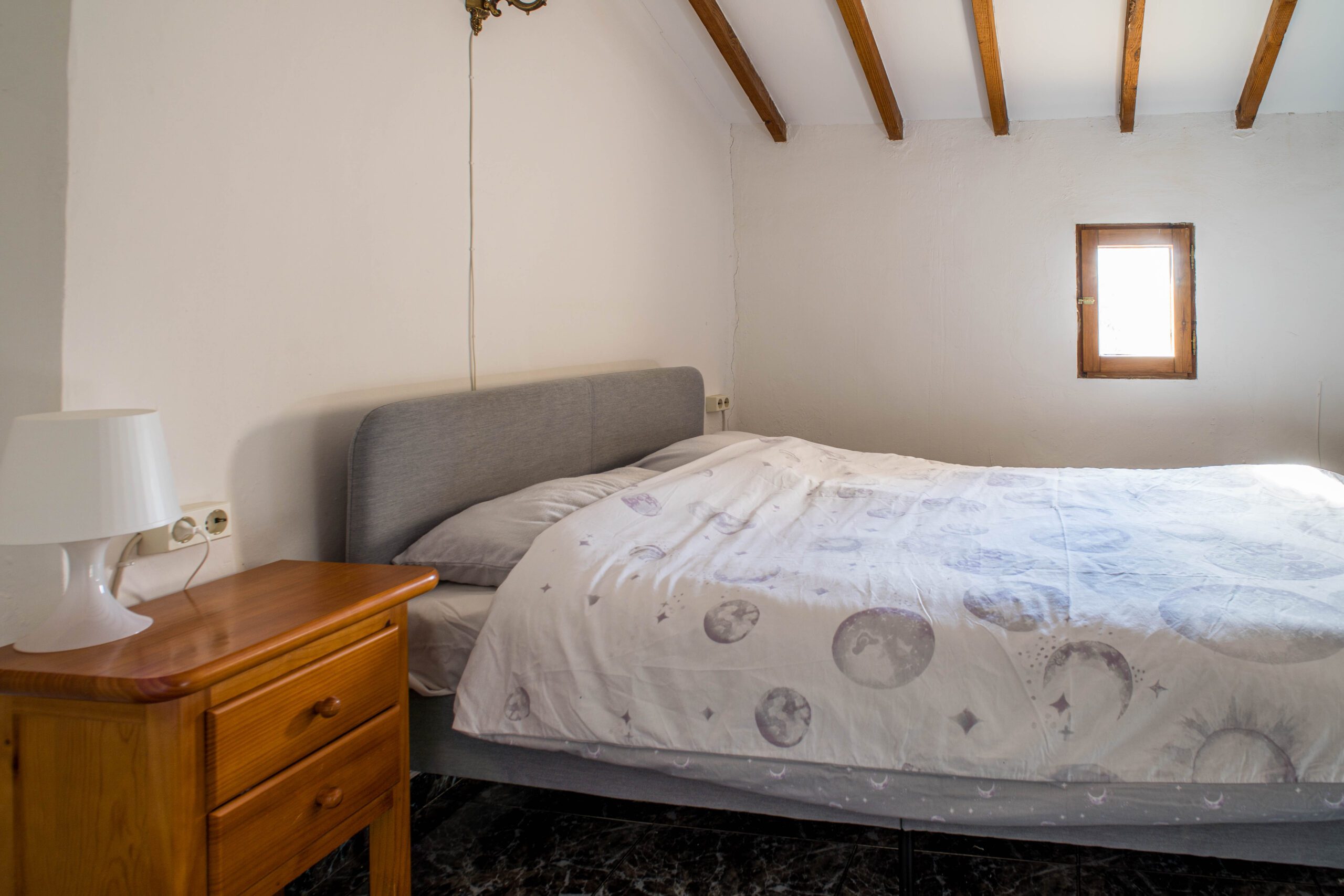 Bedroom with a bed covered in white bedding with moon patterns, a wooden bedside table with a white lamp, small window with wooden frame, and wooden ceiling beams.