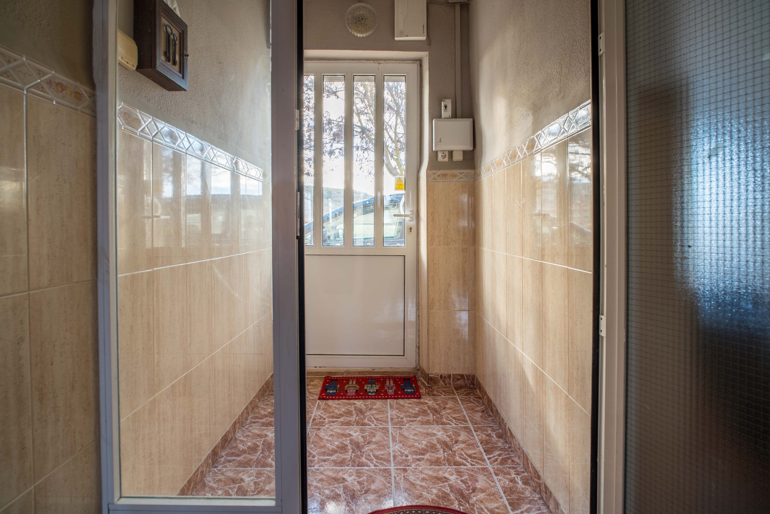 A narrow hallway with beige tiled walls and brown tiled floor, a glass door at the end with a view of a tree and a car outside.