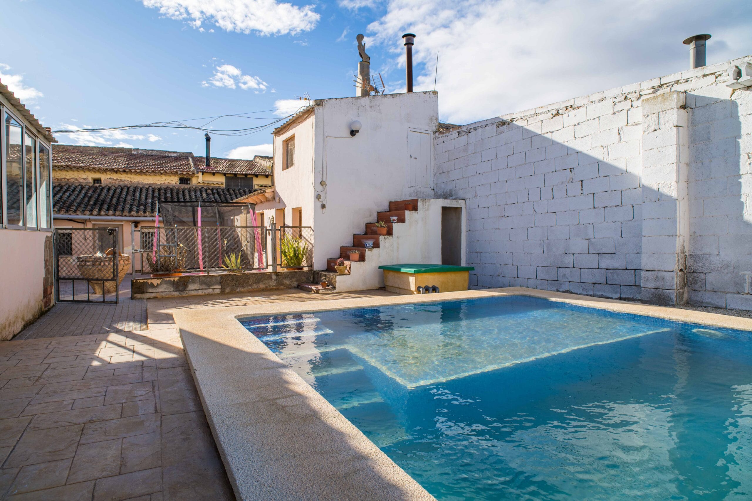 Outdoor swimming pool with clear water next to a white building with stairs and a tiled terrace under a partly cloudy blue sky.