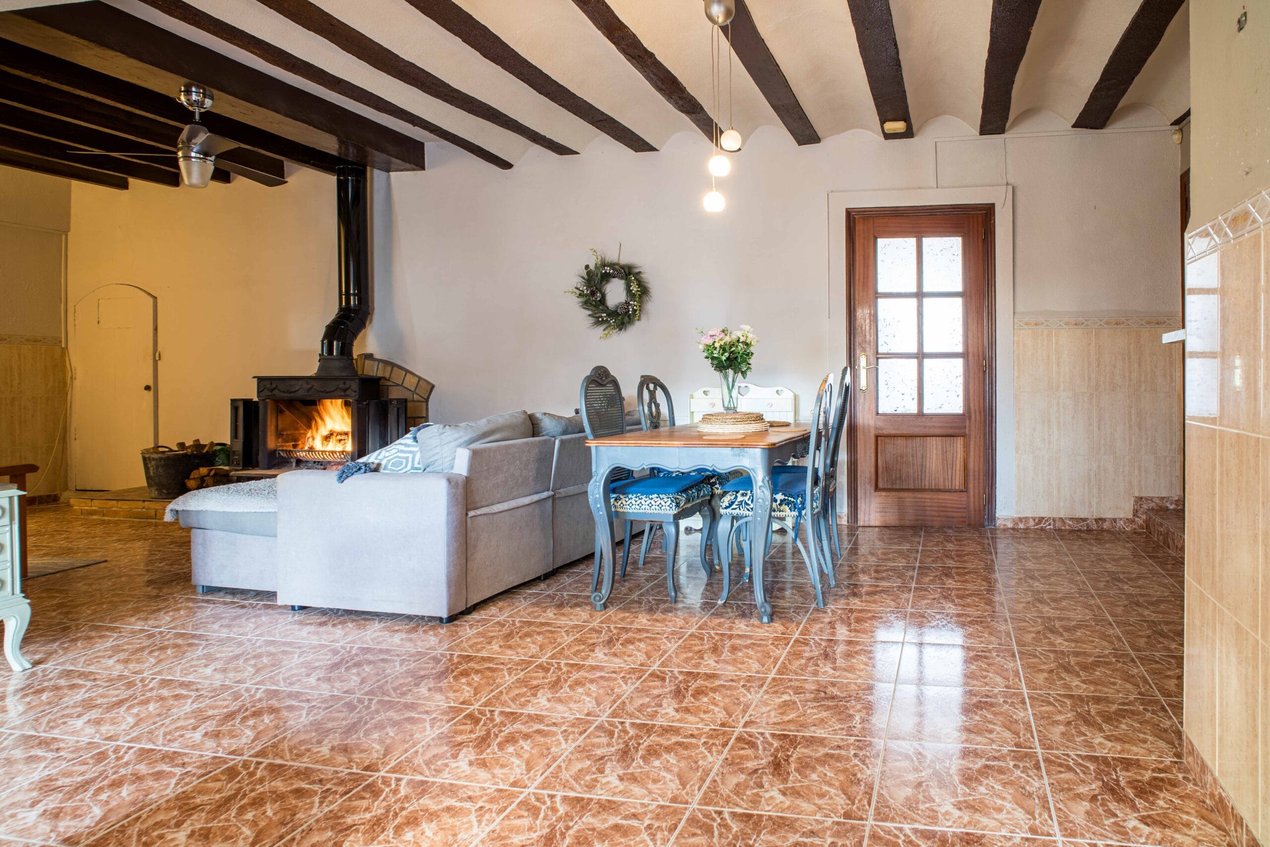 Interior view of a cozy living and dining area with a lit wood-burning fireplace, wooden ceiling beams, a gray sofa, and a wooden dining table with blue cushioned chairs.