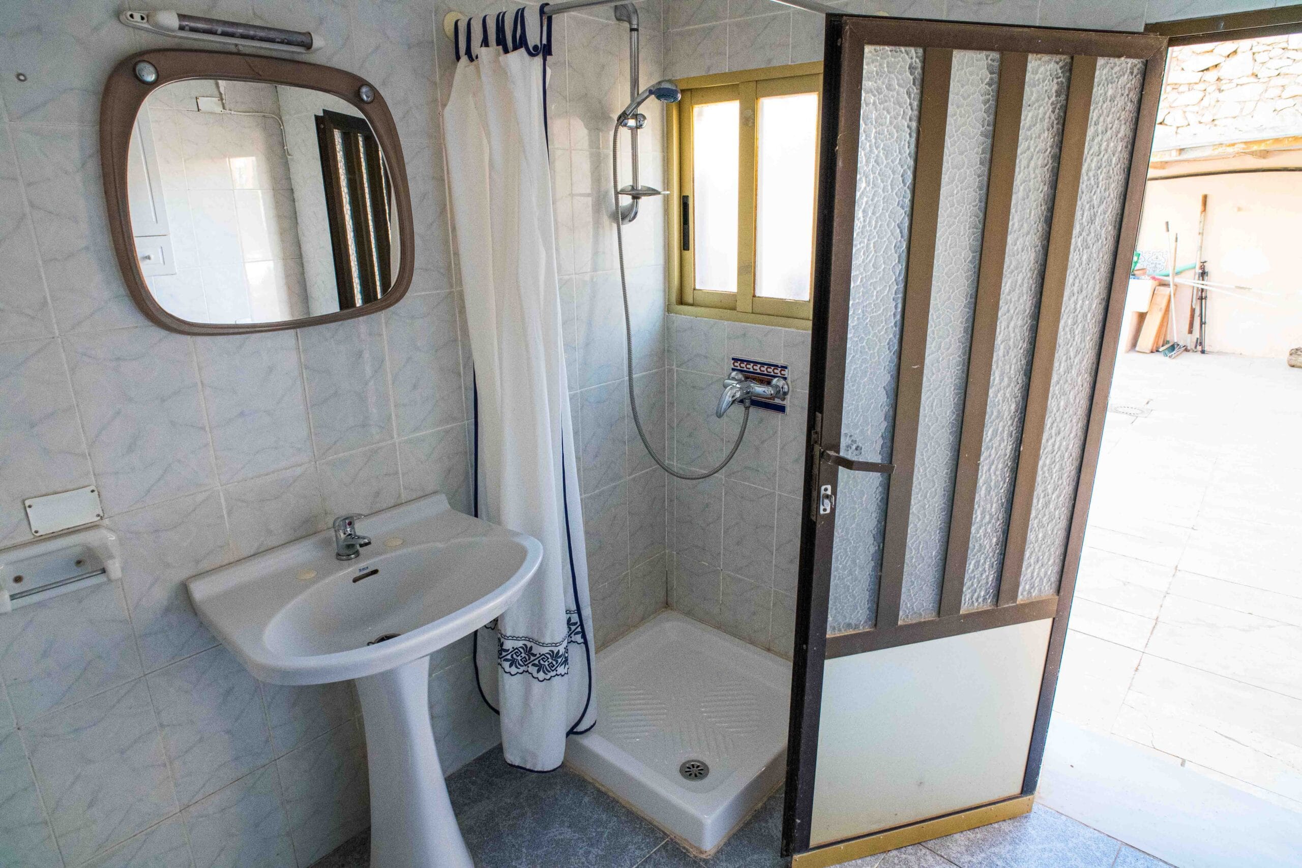 Small bathroom with a white pedestal sink, a shower with a curtain, a mirror above the sink, and a partially open door leading to an outdoor area.