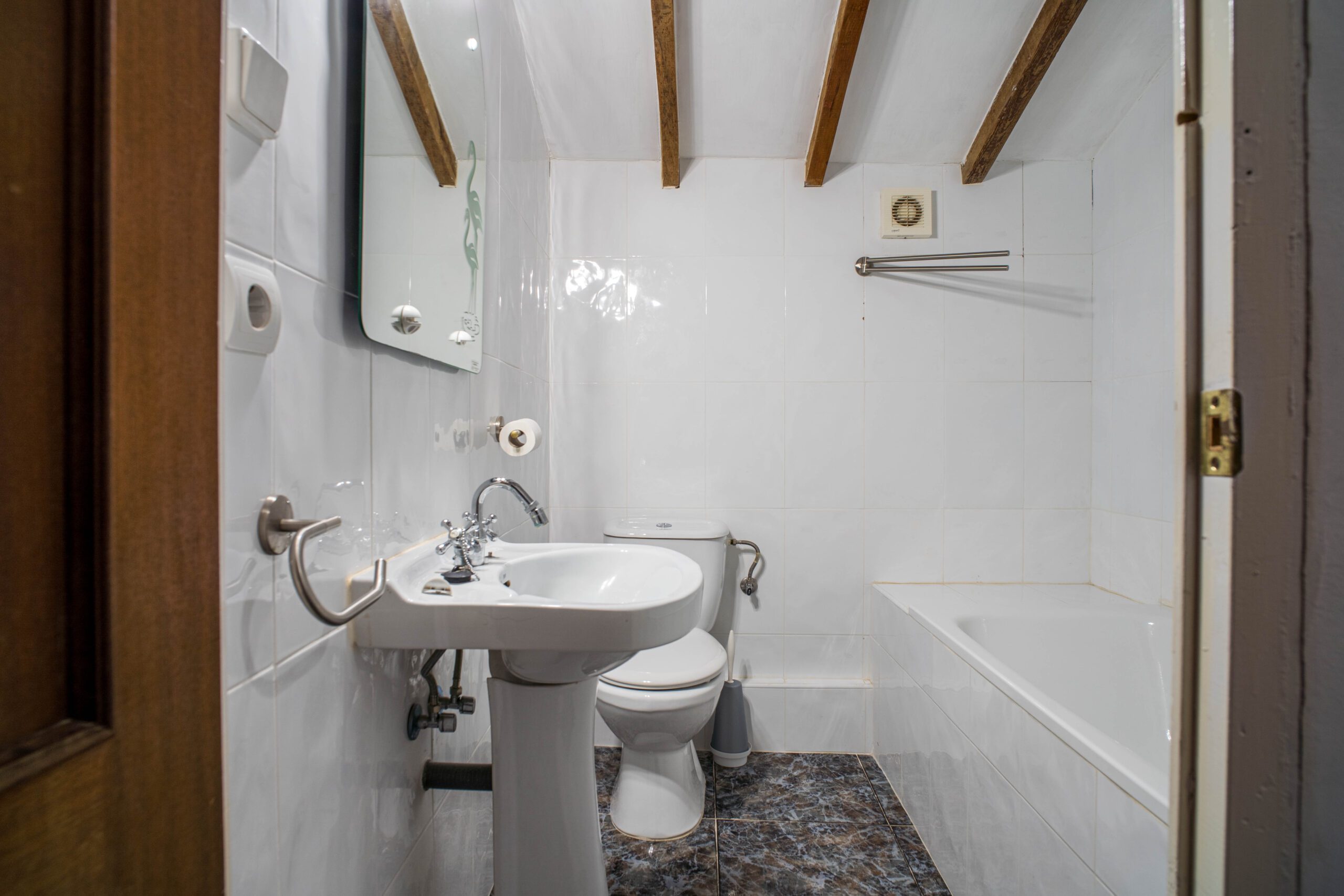 Small bathroom with white tiled walls, a bathtub on the right, a white pedestal sink with chrome faucets, a toilet, and wooden ceiling beams.