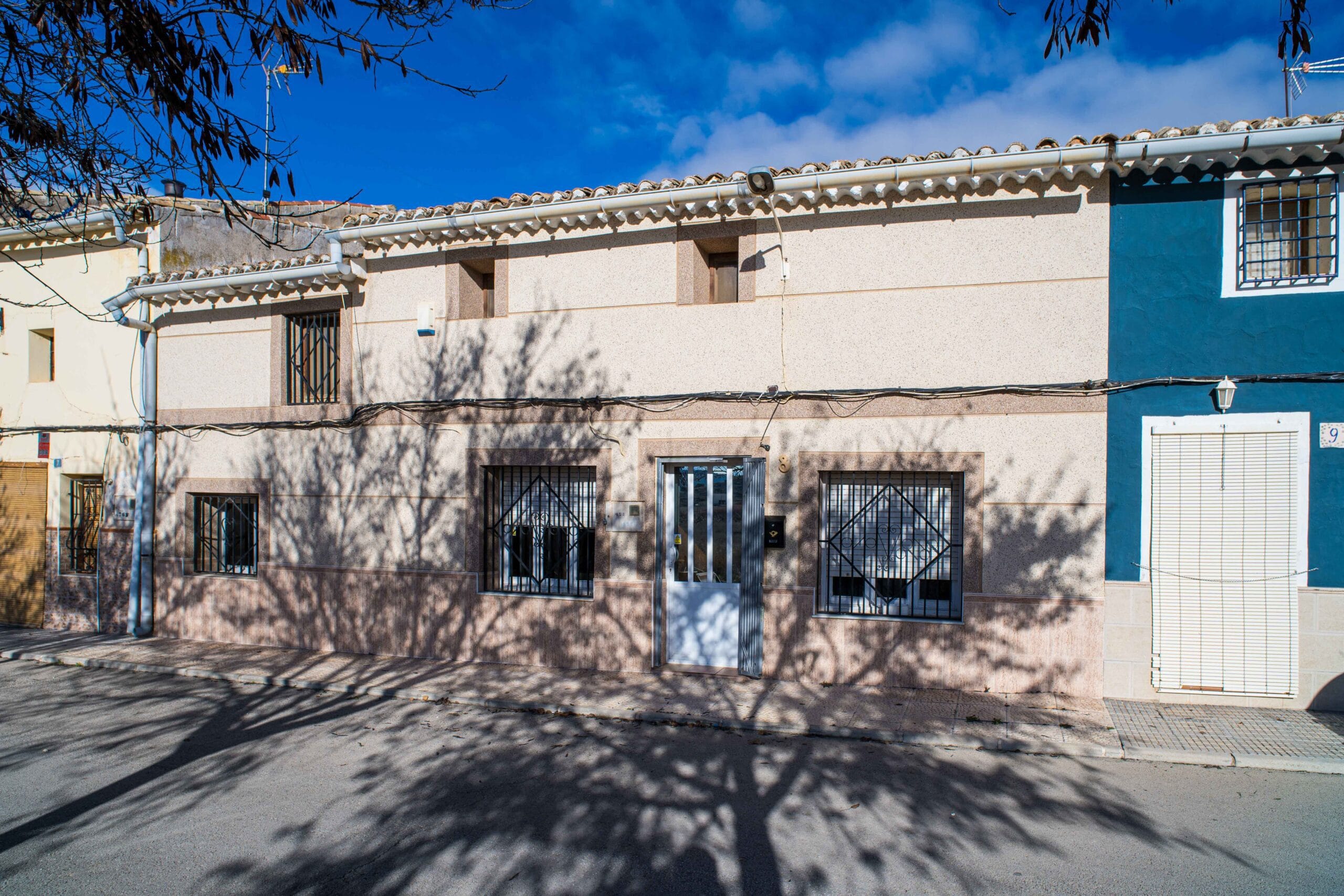 Exterior of a residential house showing barred windows, a metal gated entrance door, and shadows of tree branches on the facade and street.