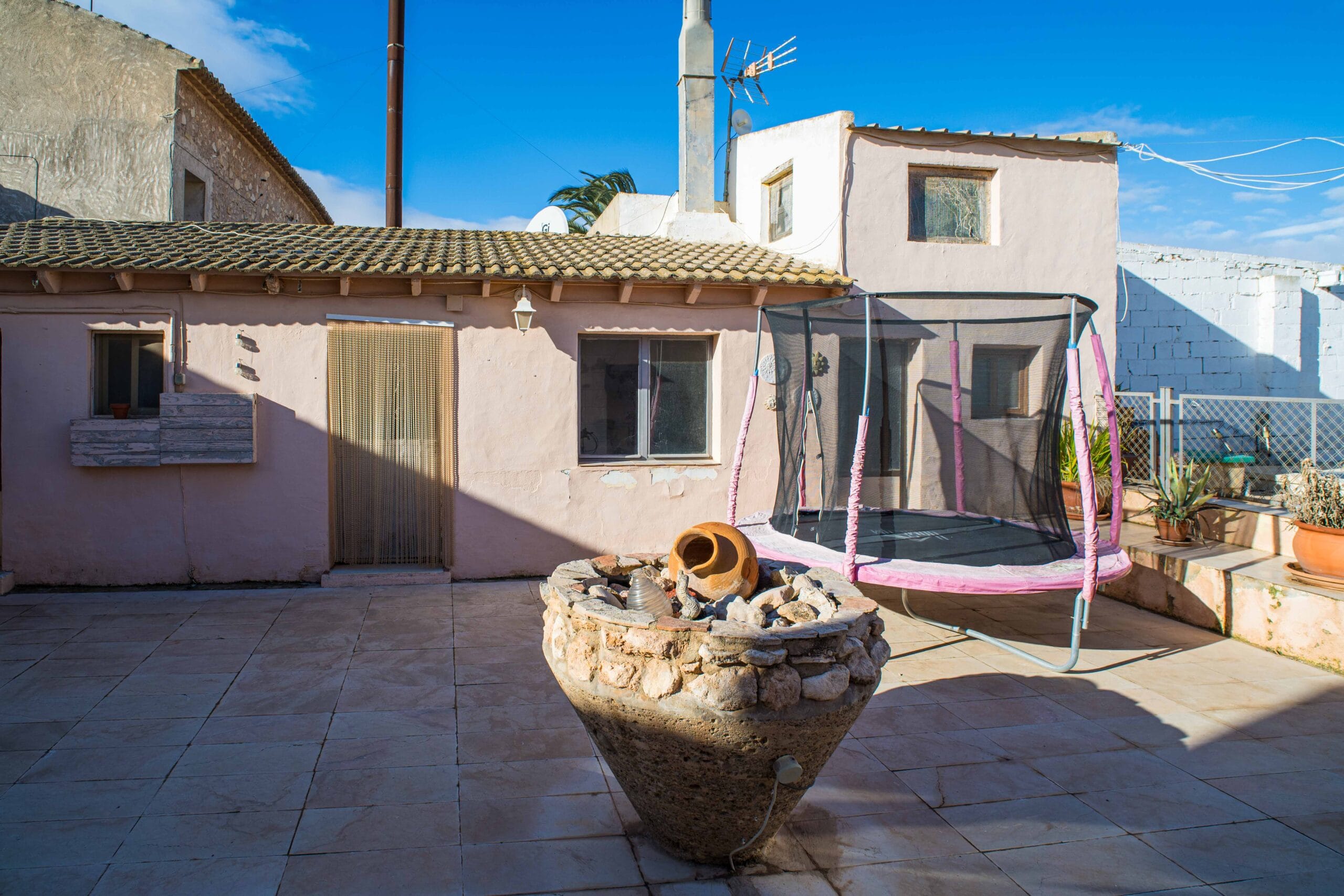Outdoor patio with tiled floor, a trampoline with pink padding, a stone planter filled with rocks and pottery, and a beige house with tiled roof under a clear blue sky.