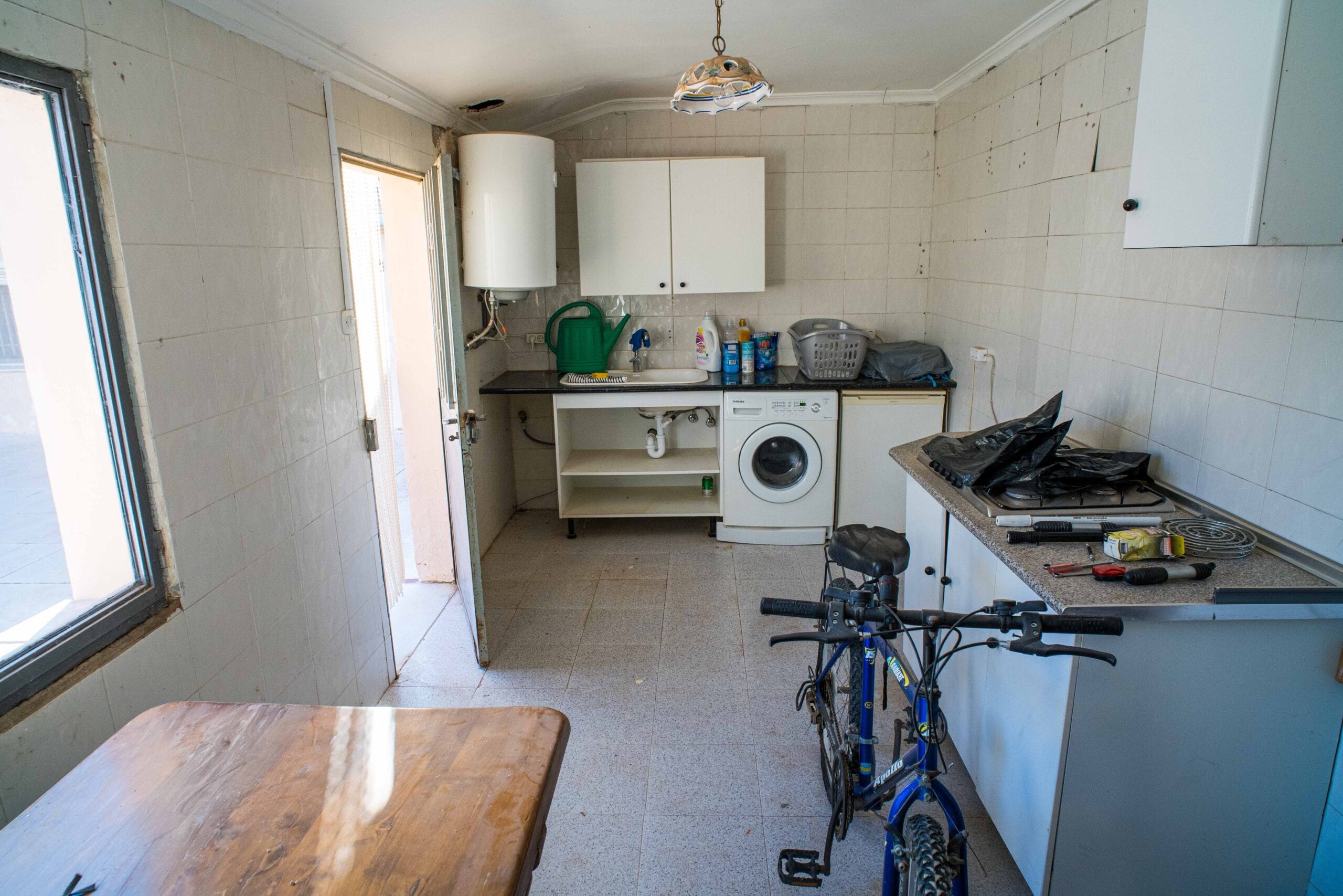 A small kitchen with white tiled walls, a washing machine under the counter, a sink, white cabinets, and a bicycle parked inside the room.