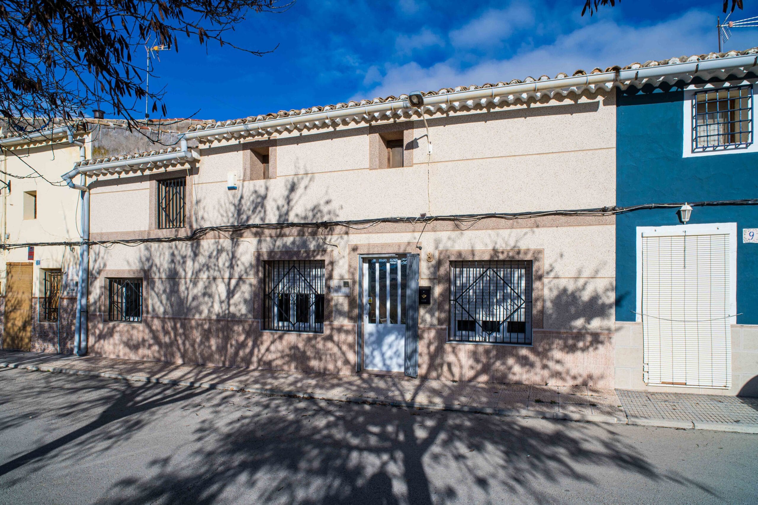 Exterior view of a residential building with barred windows and a metal gated door, shadows of tree branches on the facade, and a clear blue sky above.