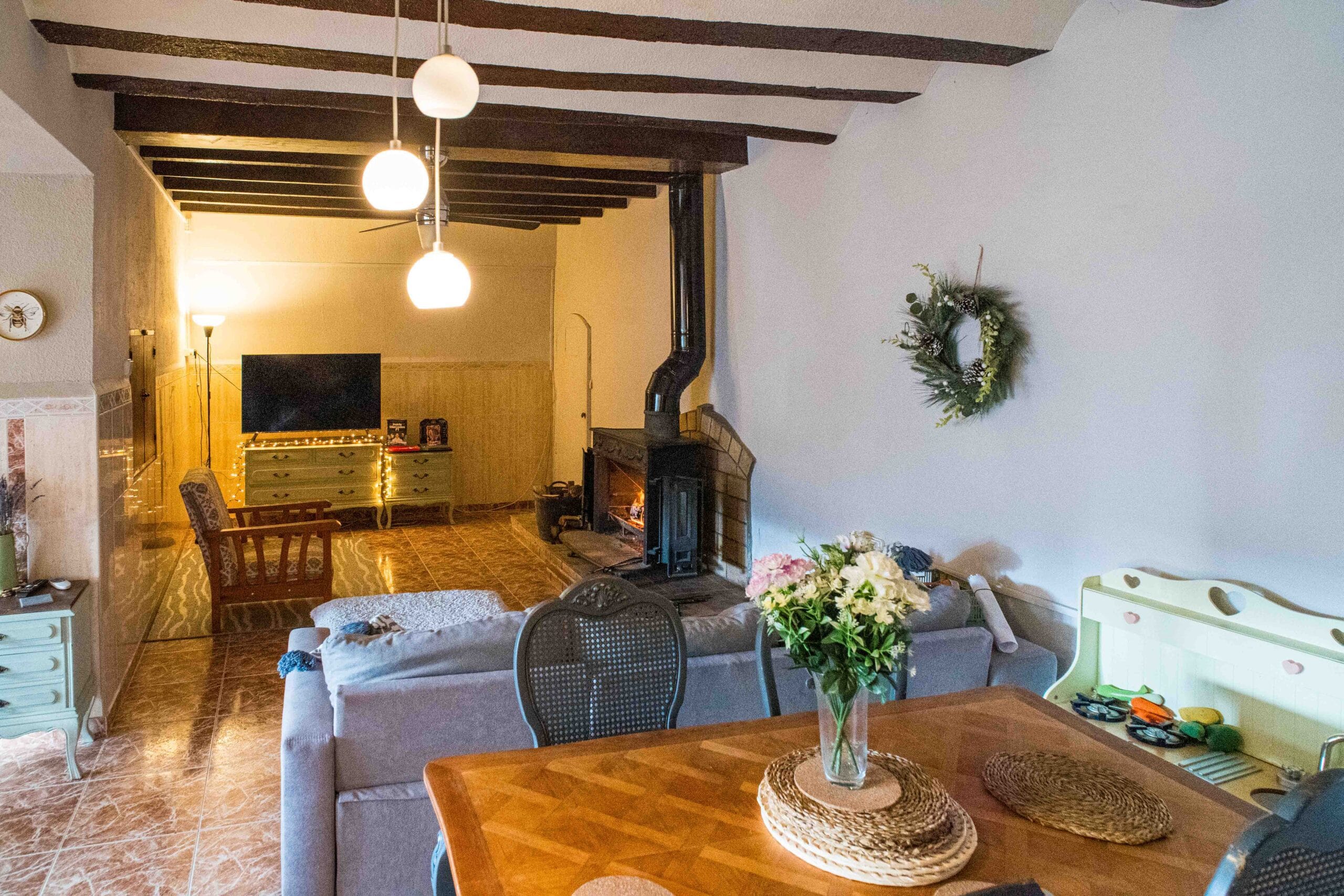 Living room with wooden beams on the ceiling, a wood-burning fireplace, a TV on a dresser, and a sofa with a dining table in the foreground.