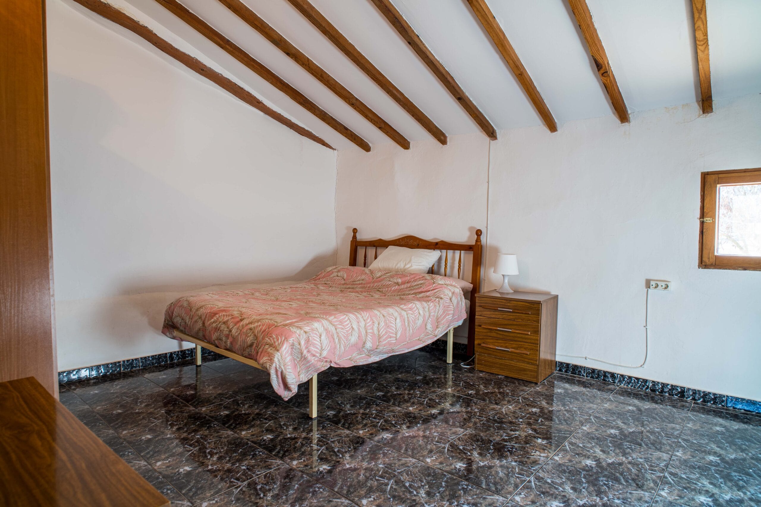 A bedroom featuring a single bed with a pink patterned bedspread, wooden headboard, a wooden bedside table with a white lamp, exposed wooden beams on the ceiling, and a small window on the right wall.