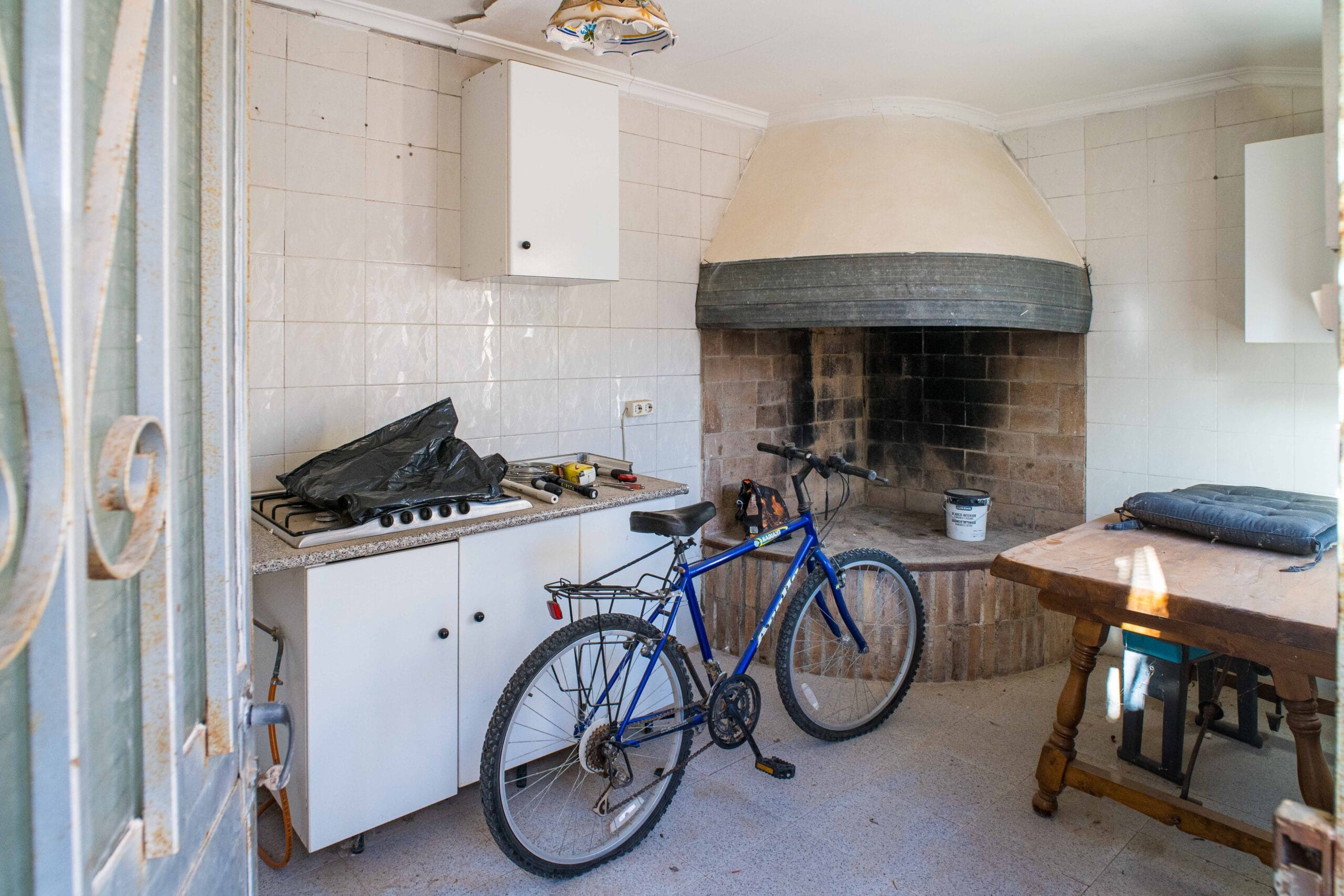 A small kitchen with white tiled walls, a large beige and black fireplace, a gas stove with a black plastic bag on top, and a blue bicycle parked inside next to a wooden table with cushions.