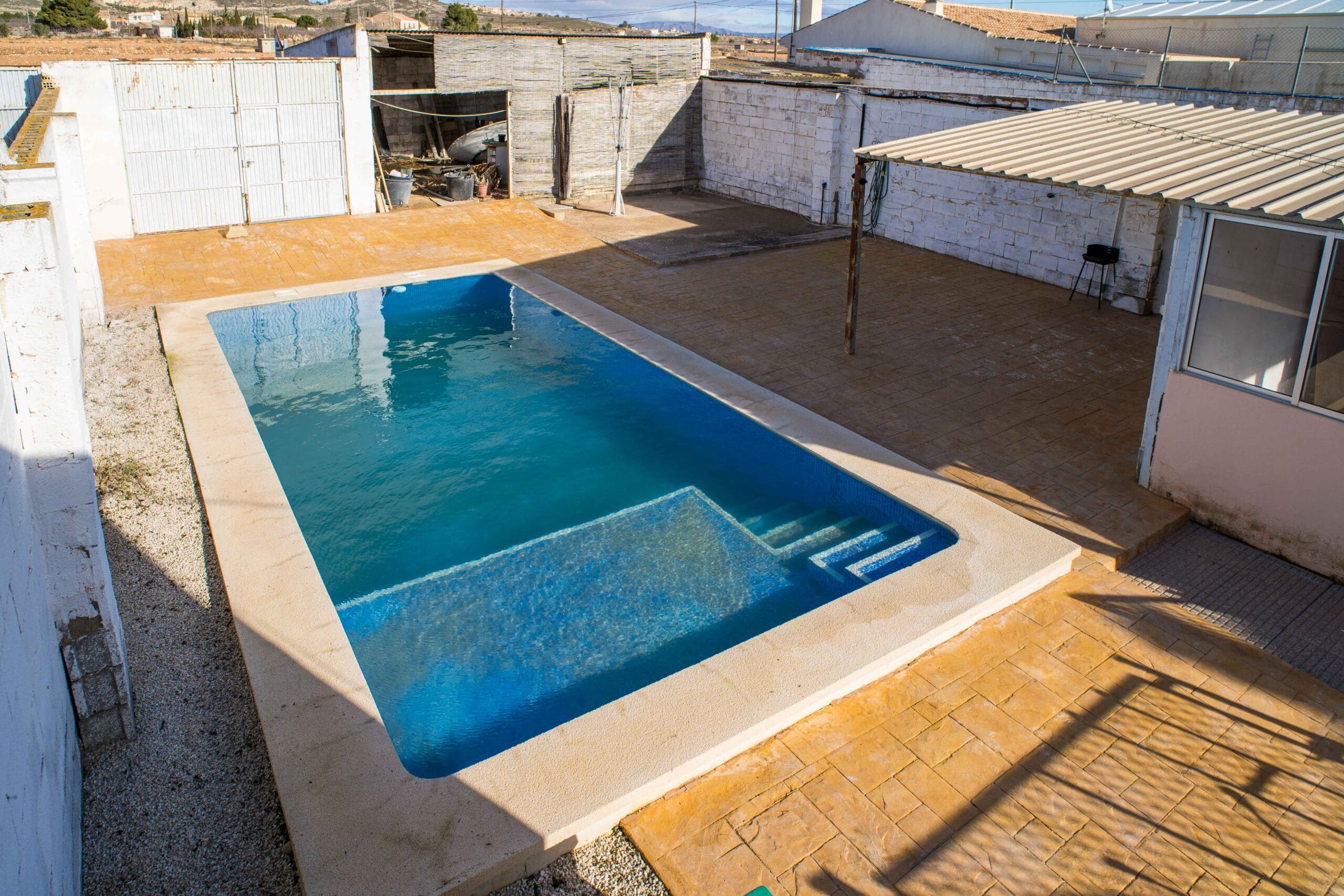 Outdoor swimming pool with clear blue water next to a tiled terrace area enclosed by white brick walls and a metal gate.
