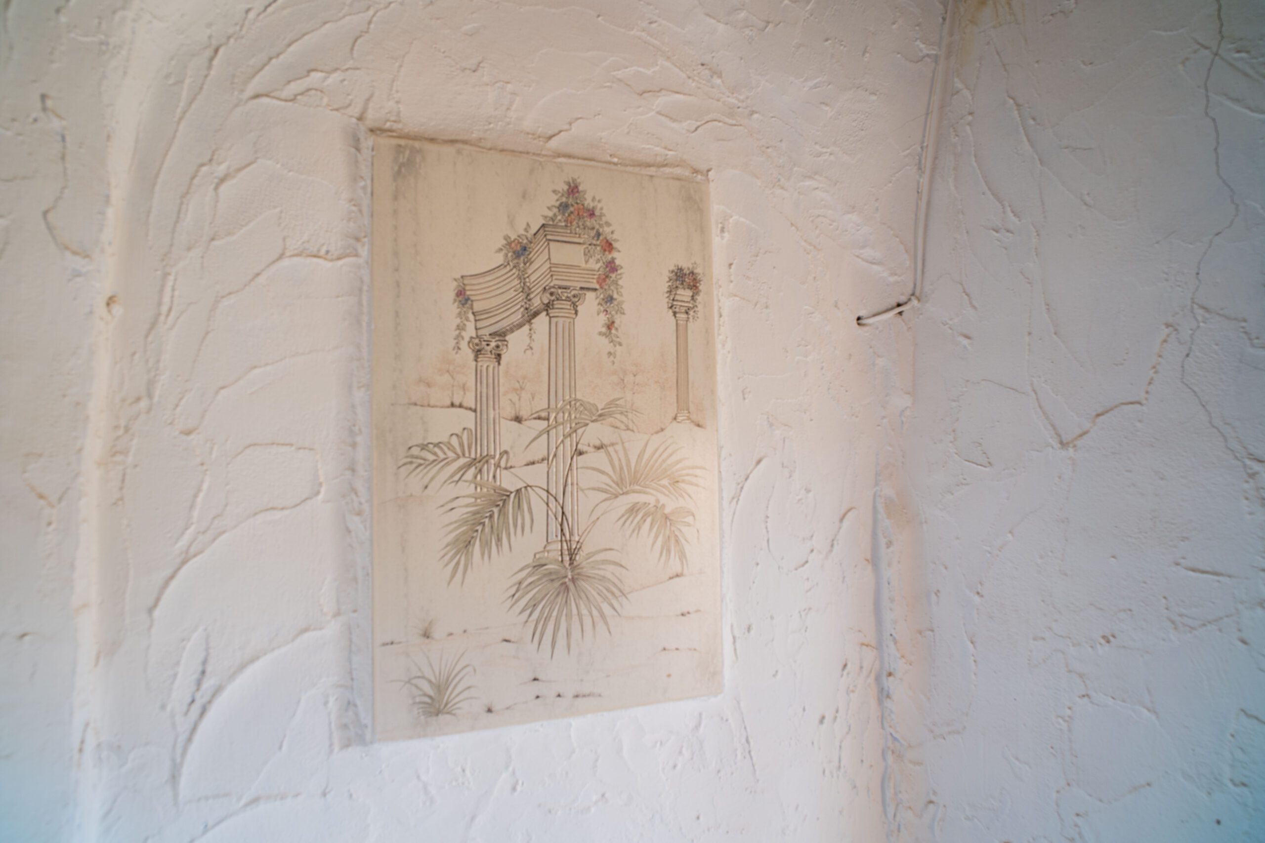 A textured white wall with a framed painting showing classical columns decorated with flowers and plants.