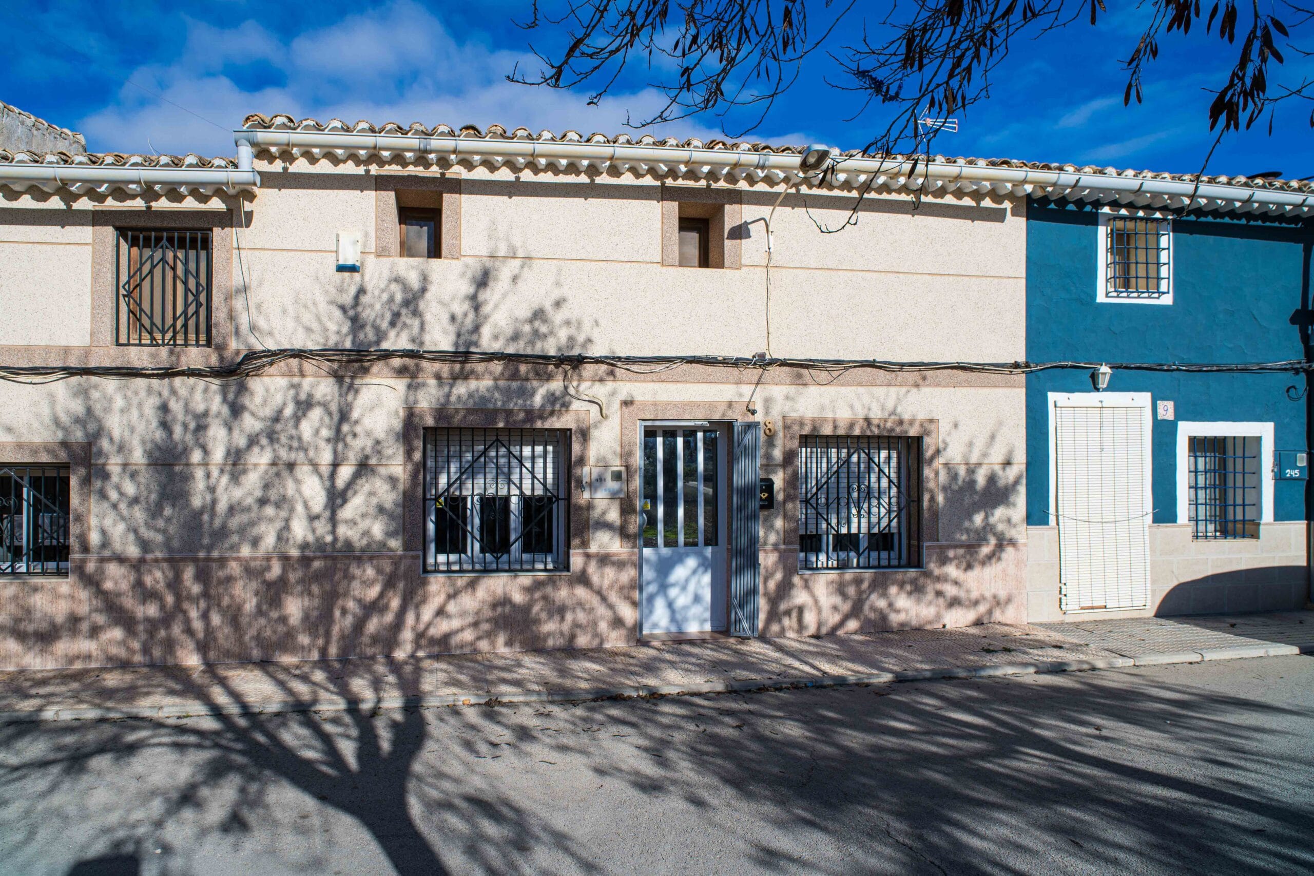 Exterior view of a residential building showing a beige and blue facade with barred windows, a gated front door, and shadows of tree branches on the wall and ground.