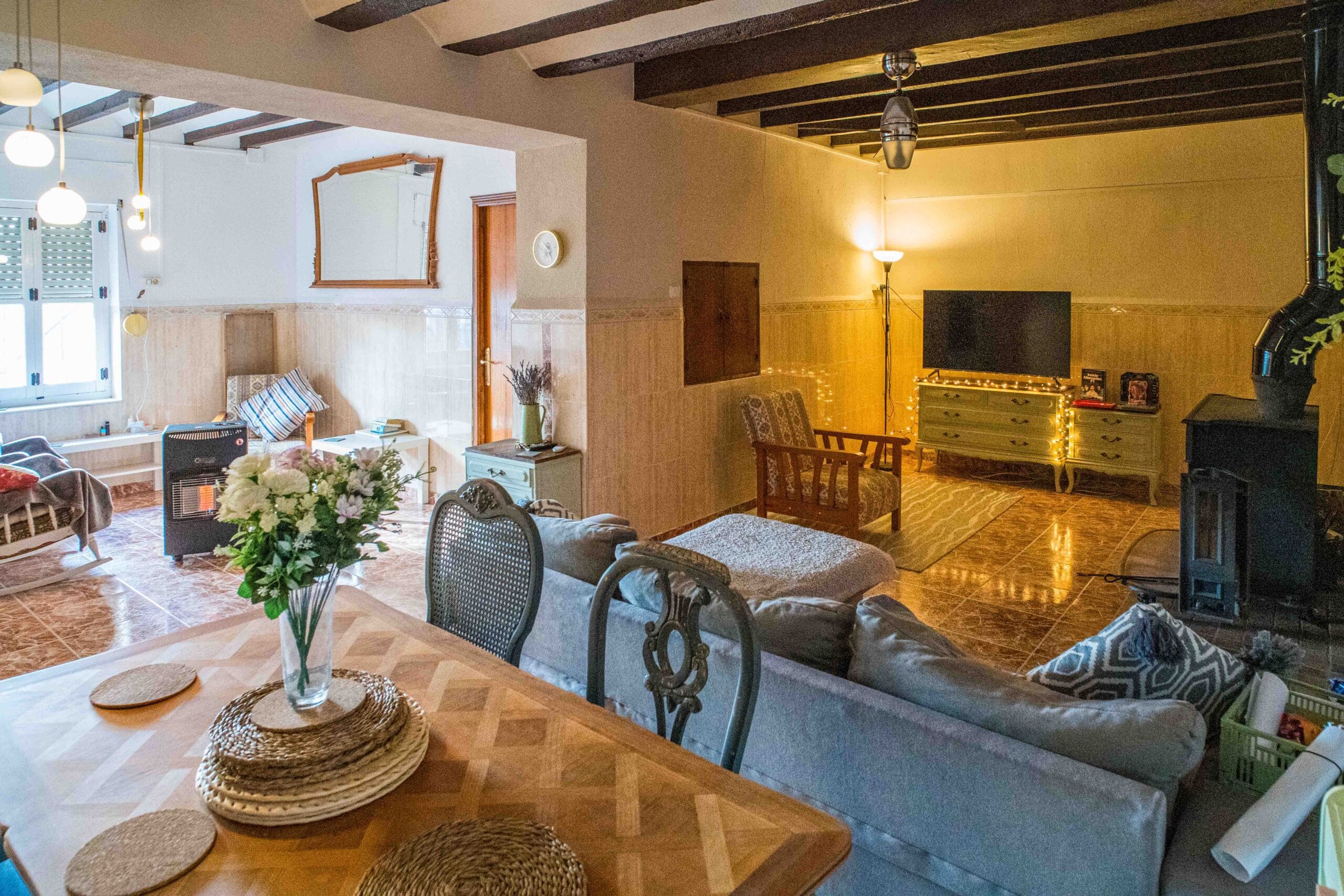 A cozy living room with exposed dark wooden ceiling beams, a fireplace, a TV on a dresser, armchairs, a sofa, and a wooden dining table with placemats and a vase of flowers.