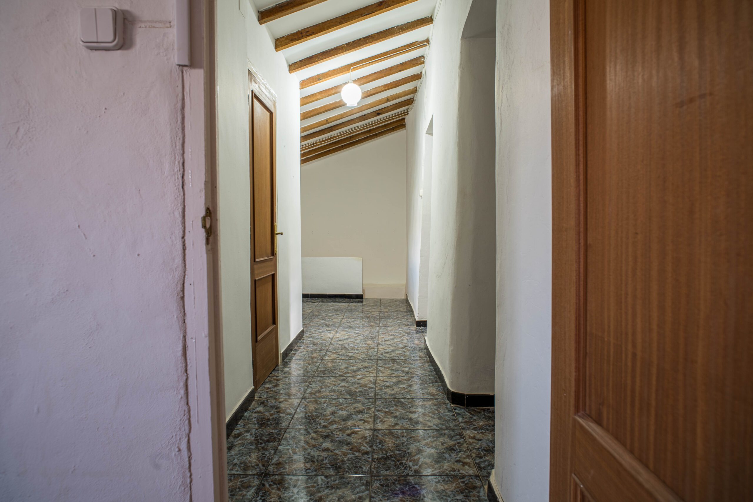 A narrow hallway with tiled floor, white walls, wooden doors on the sides, and a sloped ceiling with exposed wooden beams and a hanging light fixture.