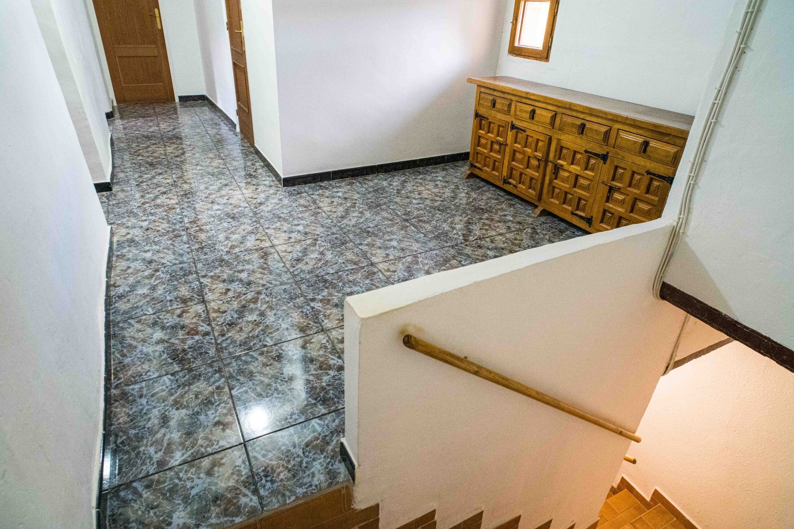Interior hallway featuring marble-patterned tile floor, wooden doors, a wooden sideboard, and a staircase with wooden handrail going down.
