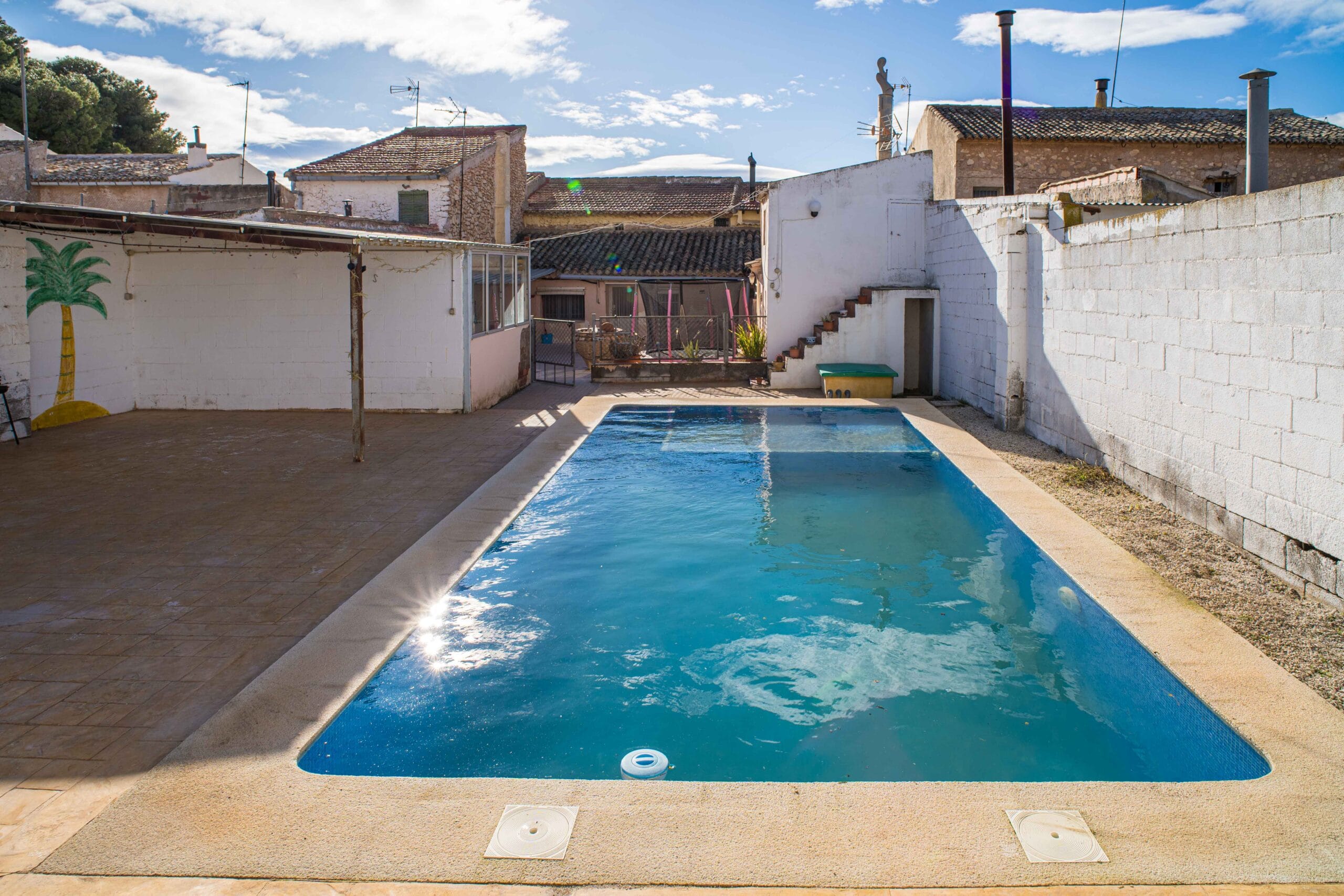 Outdoor swimming pool with clear blue water, tiled terrace area, white walls enclosing the space, and rustic houses with tiled roofs in the background under a blue sky with scattered clouds.