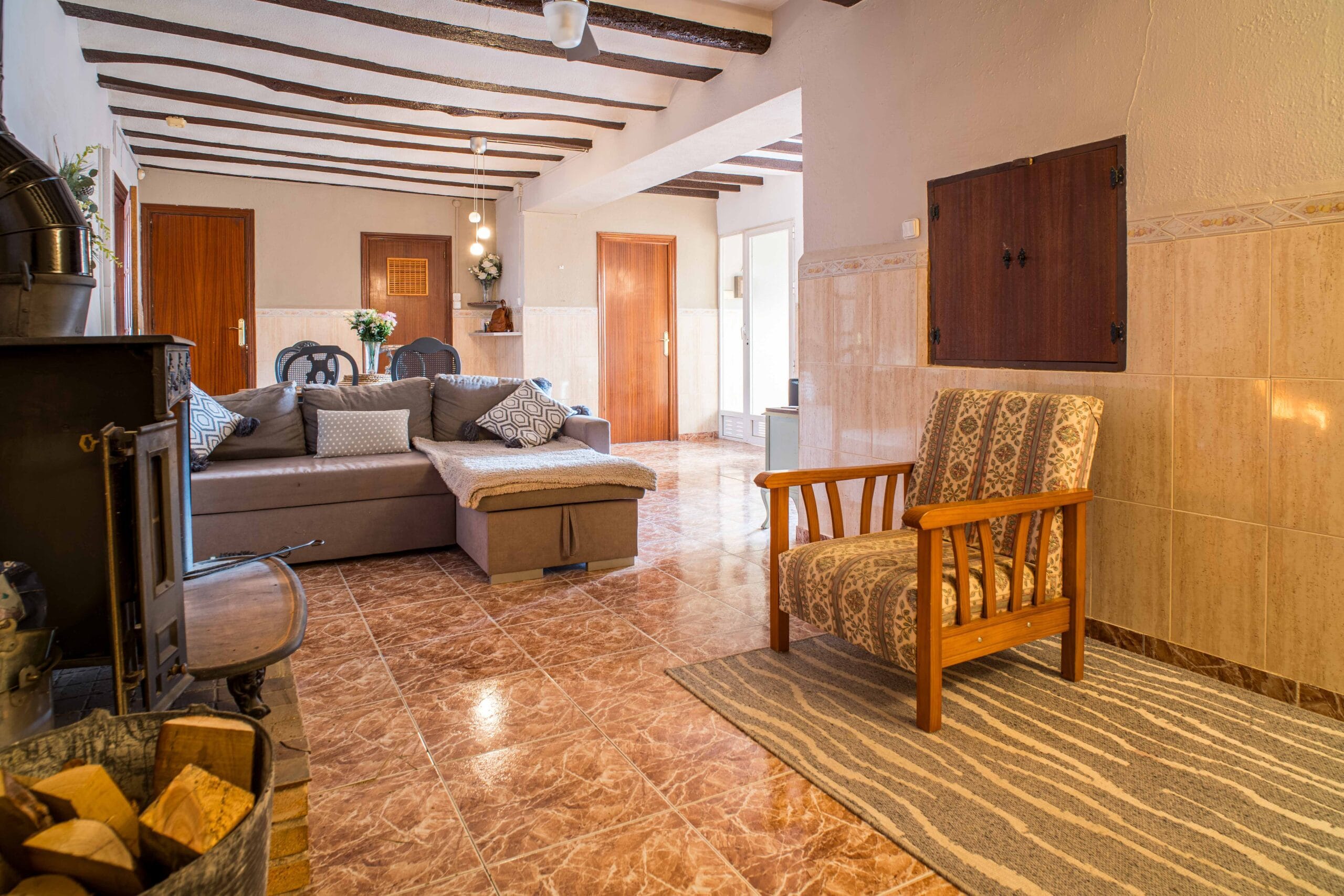 Living room with a wood-burning fireplace, a gray sectional sofa with patterned cushions, wooden ceiling beams, and a wooden armchair on a striped rug.