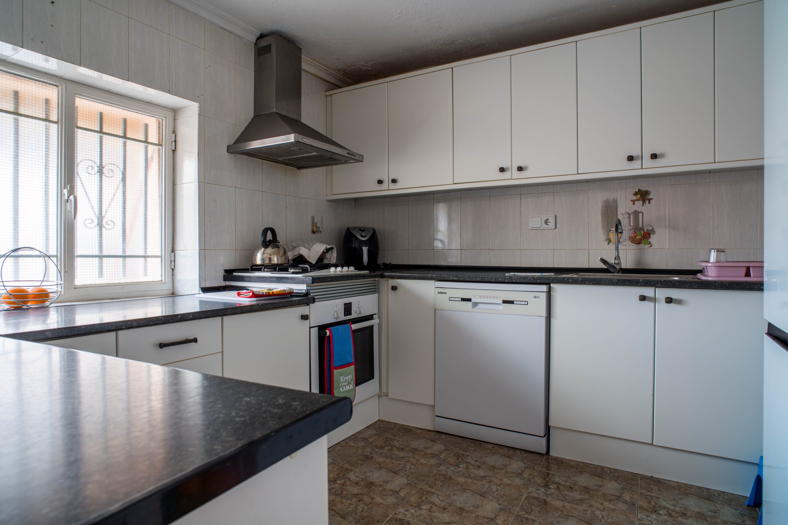 Compact kitchen featuring white cabinets, black countertops, a stove with a kettle, a dishwasher, and a window with metal bars.
