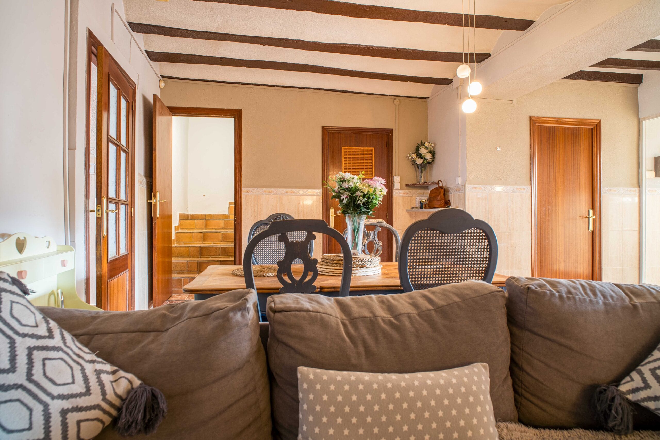 Dining room featuring a wooden table with chairs, a vase of flowers, exposed ceiling beams, and wooden doors with stairs visible through one doorway.