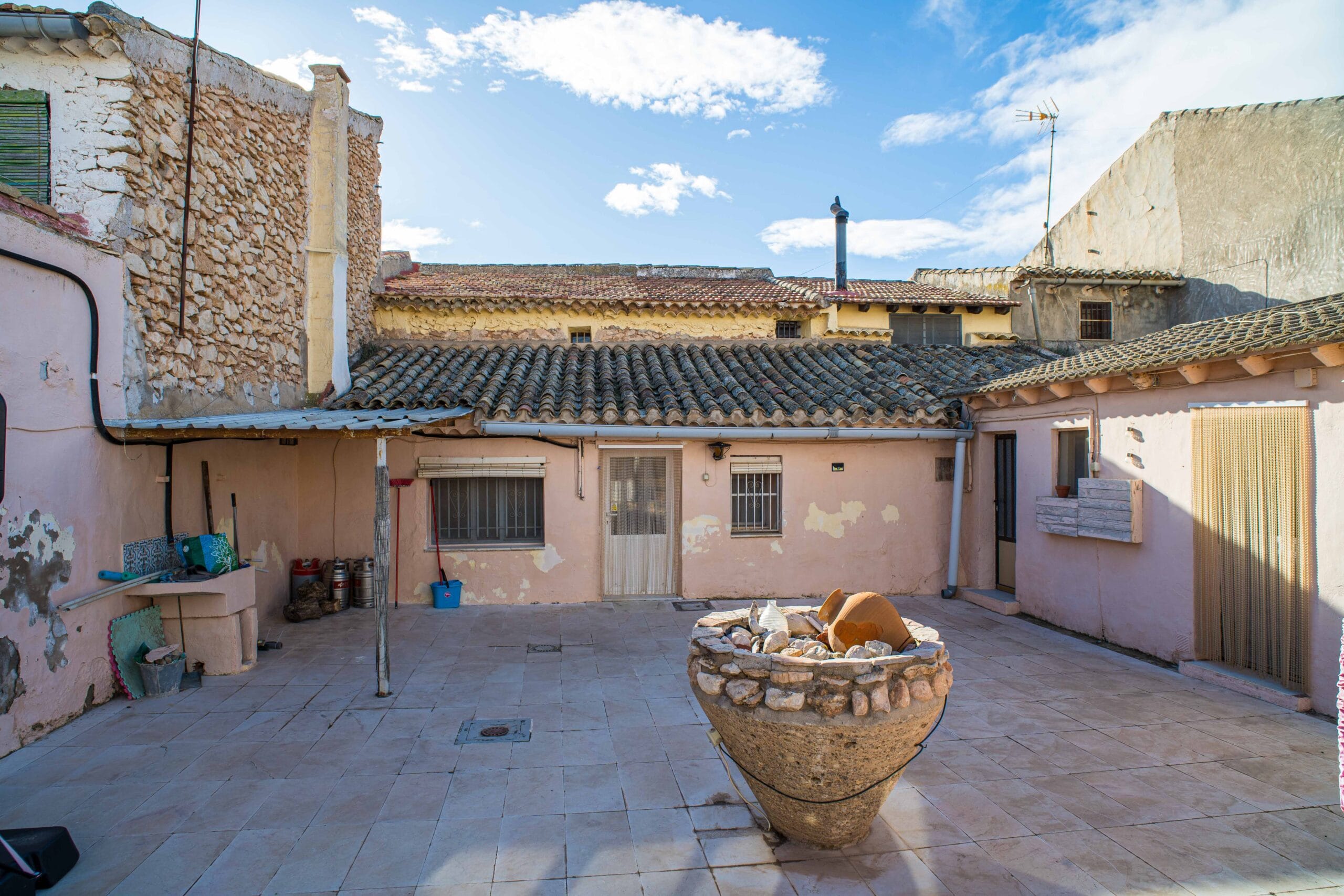 Outdoor courtyard area featuring tiled floor, stone walls, a large stone planter filled with rocks and pottery, and a clear blue sky with some clouds.