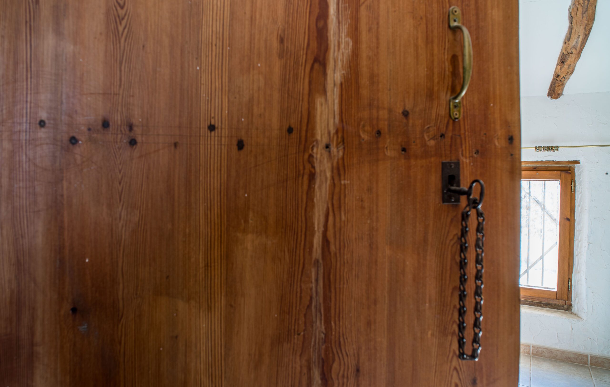 A wooden door with visible grain and knots, featuring a brass handle and a chain lock, partially open showing a window with bars and tiled floor inside.