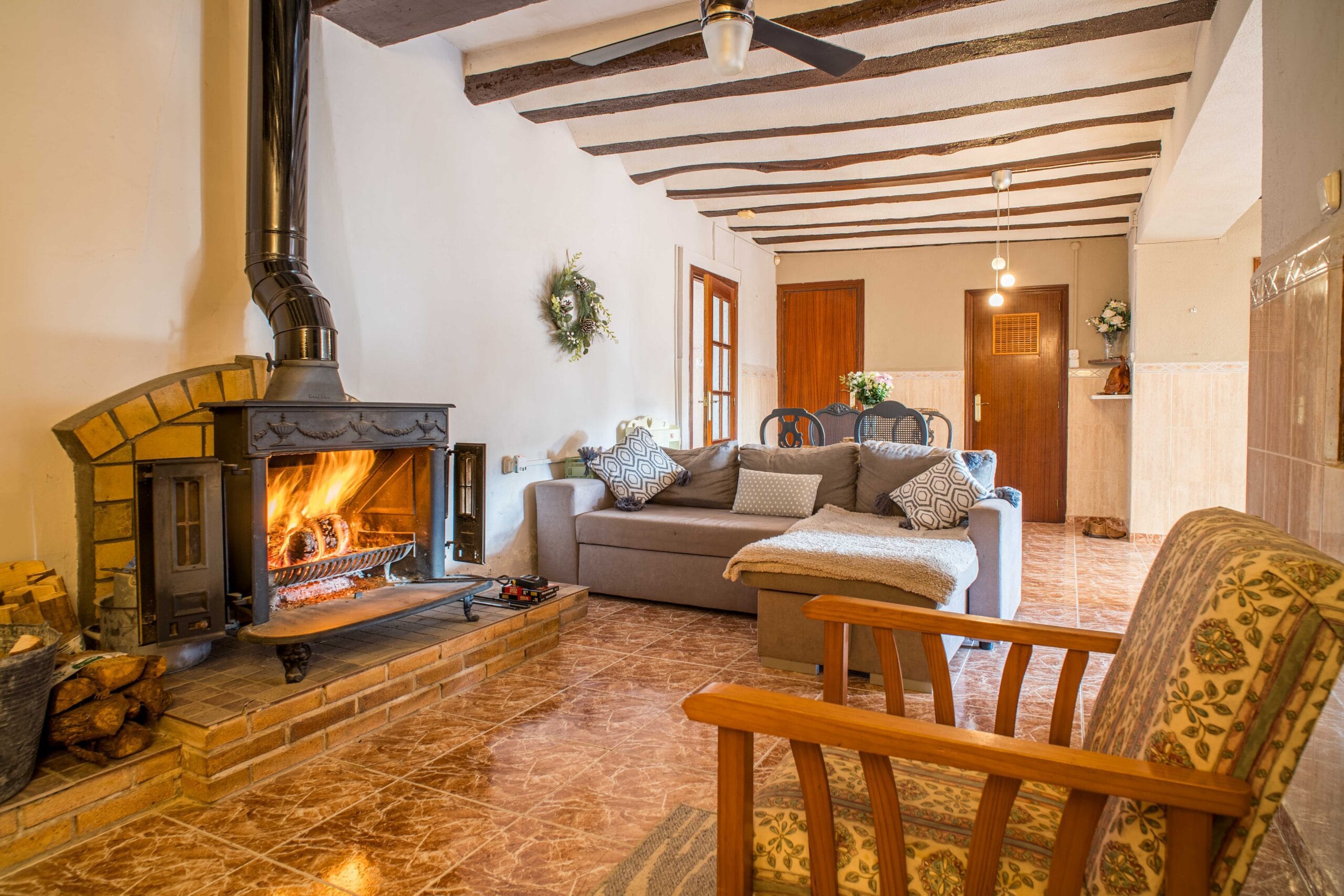 Living room with a burning fireplace, a gray sofa with patterned cushions, wooden ceiling beams, and tiled floor.