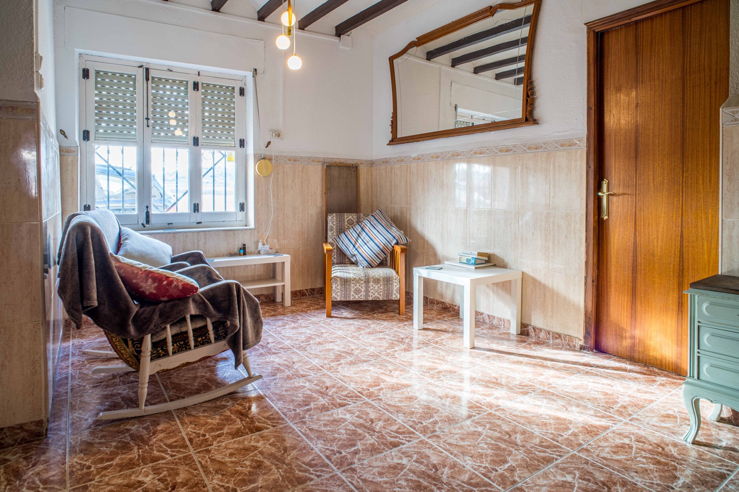 Small sitting area with two chairs, a large wooden-framed mirror on the wall, tiled floor, and a wooden door.