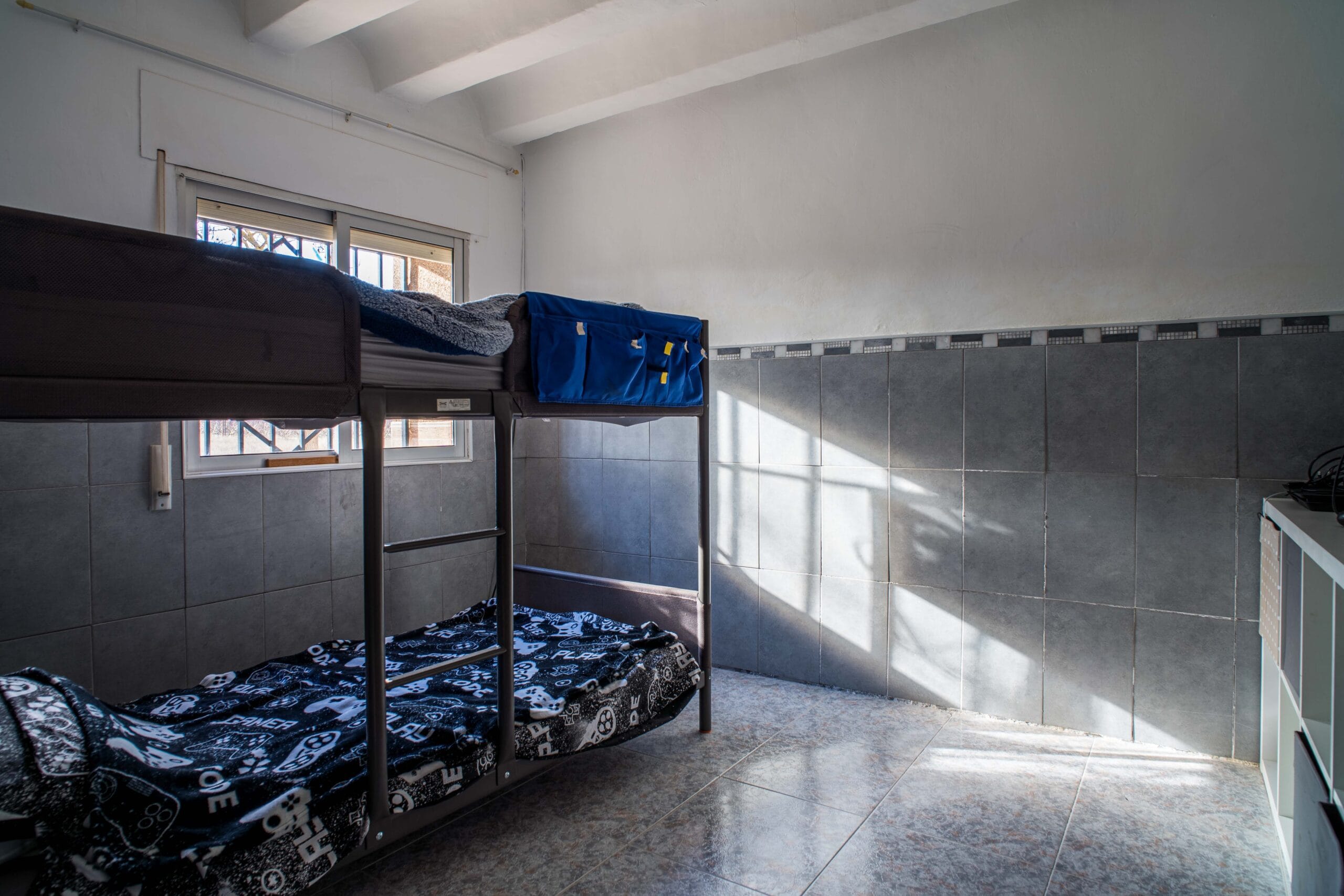 A small bedroom with a metal bunk bed covered in black and white patterned bedding, tiled floor, and sunlight streaming through a window with bars.