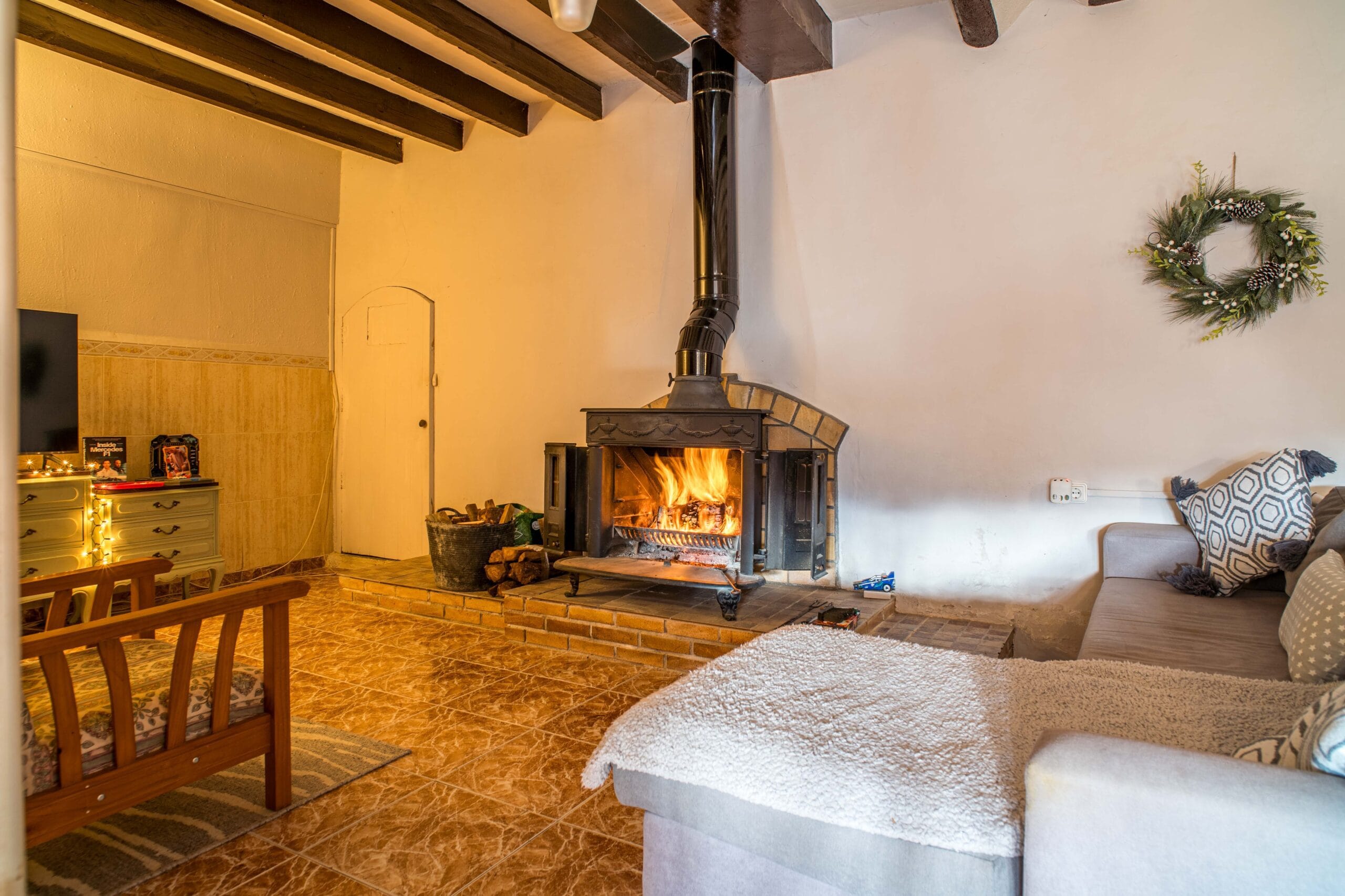 Living room with a lit fireplace, wooden ceiling beams, a sofa with patterned cushions, and a wooden chair on tiled floor.