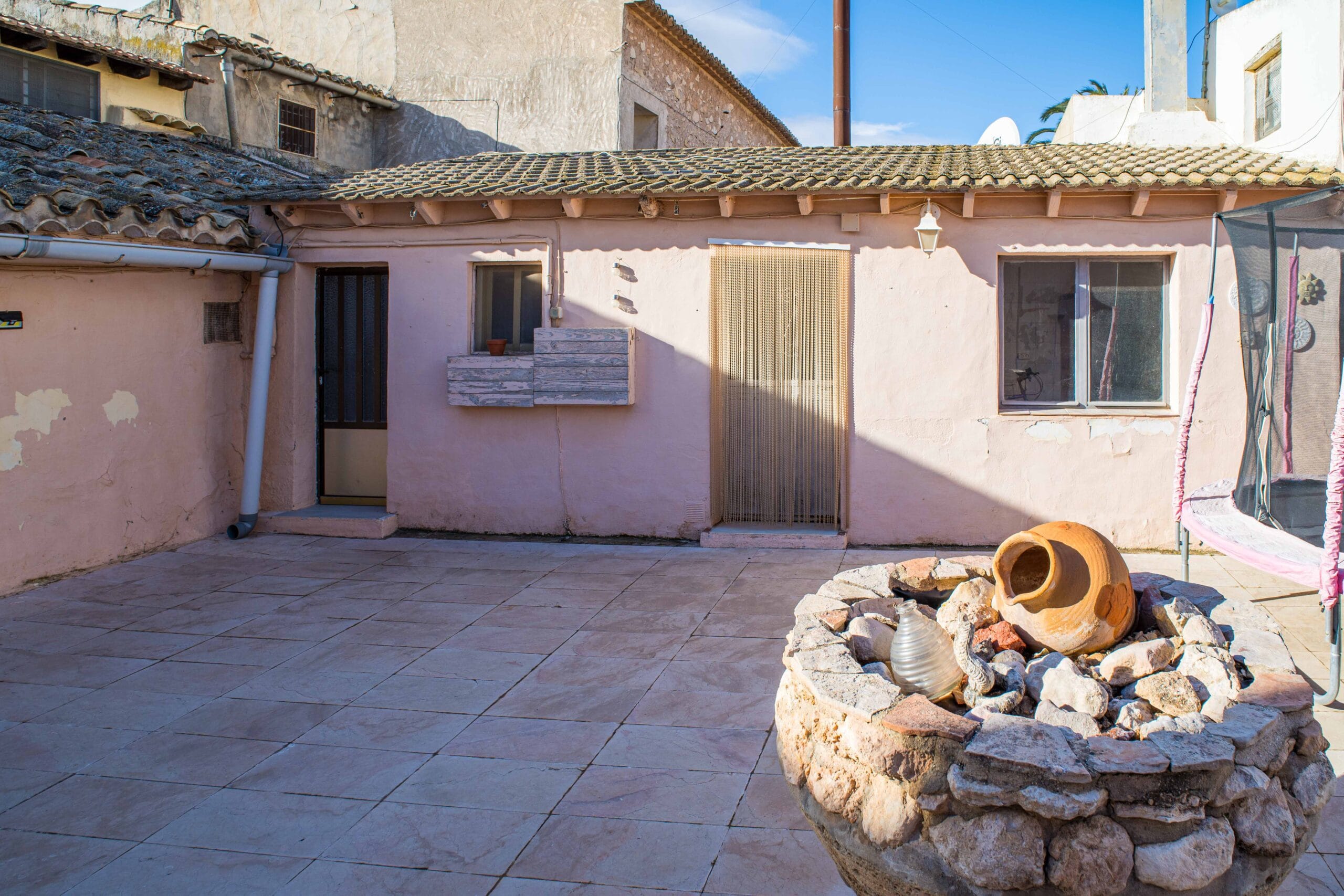 Outdoor courtyard area with tiled floor, beige walls, a small window, two doors, and a stone planter filled with rocks and pottery under clear blue sky.
