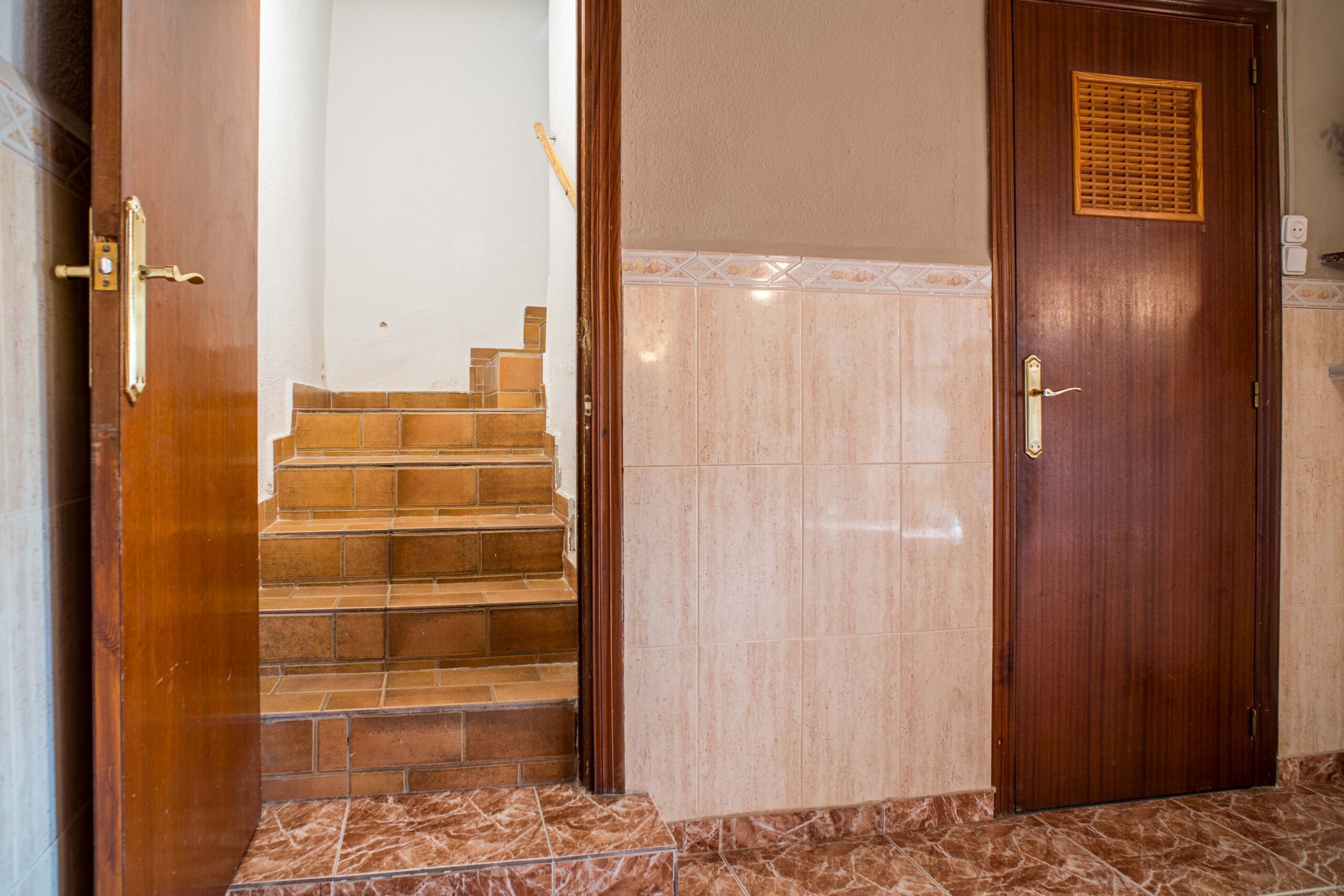 A hallway with brown tiled stairs leading upwards, brown wooden doors on either side, and beige tiled walls.