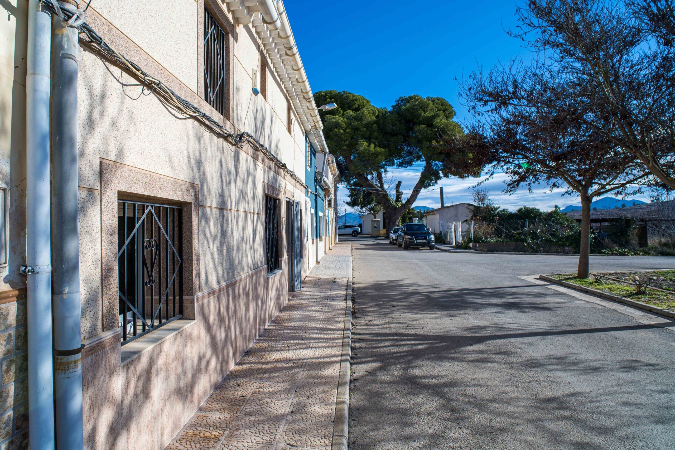 A street with residential buildings on the left, parked cars, and trees on the right under a bright blue sky.