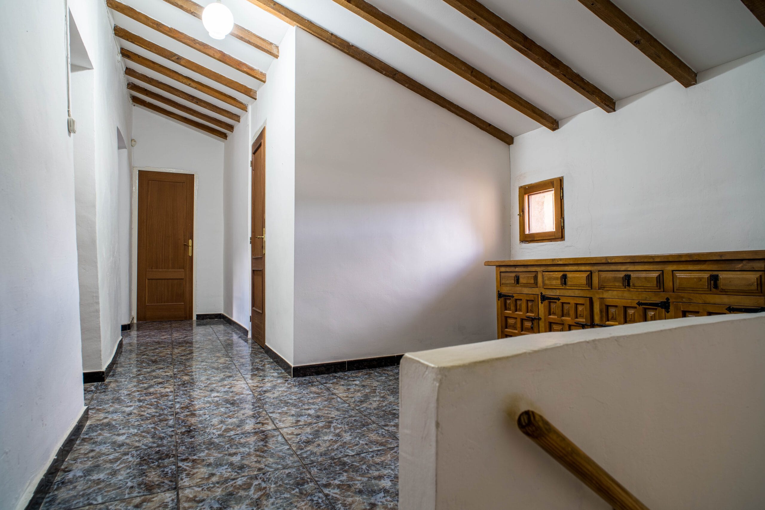 Interior hallway with white walls, exposed wooden ceiling beams, tiled floor, wooden doors, and a small wooden window.
